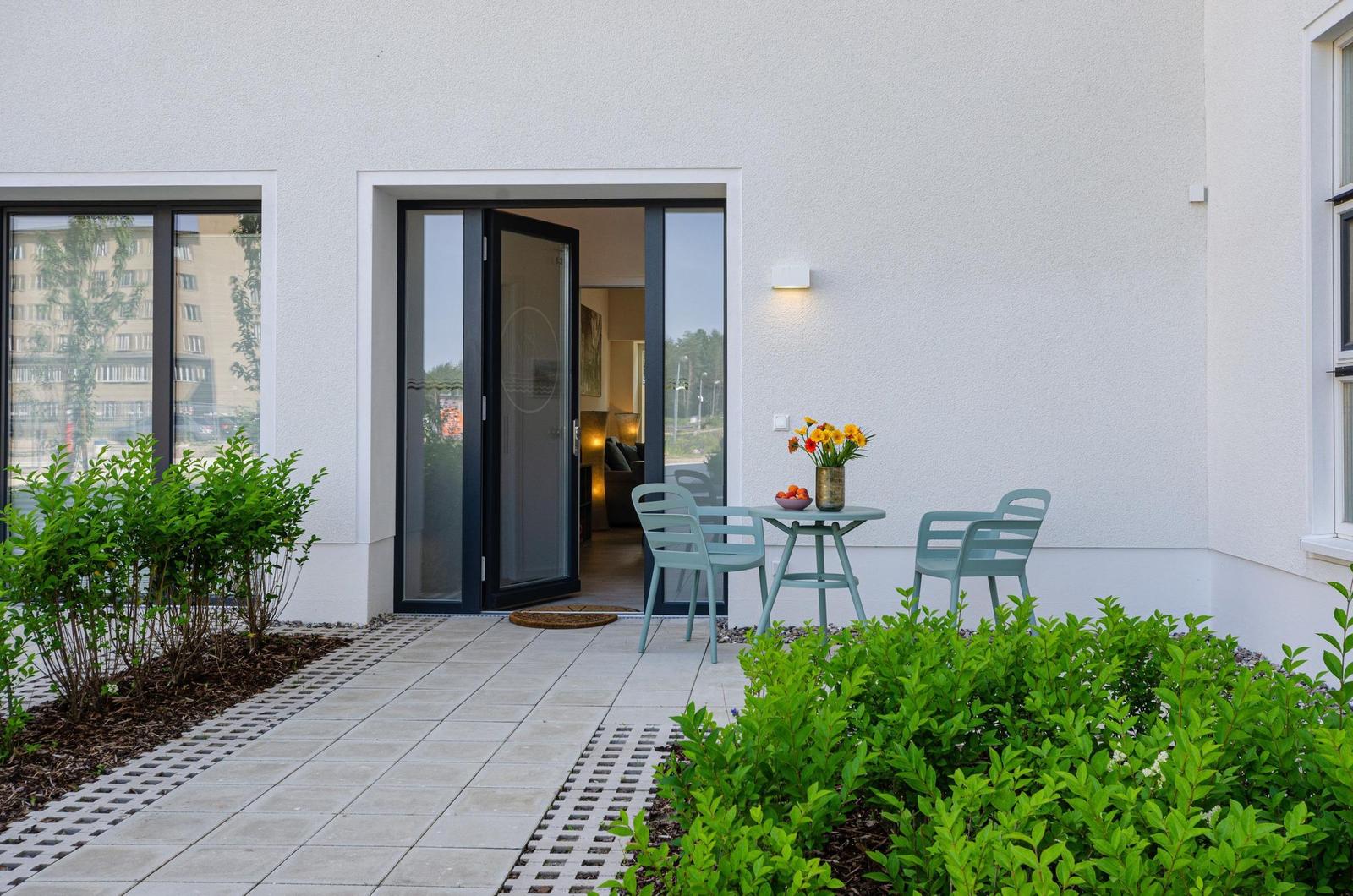Terrasse mit Tisch, zwei Stühlen und Blumen vor einer Glastür.