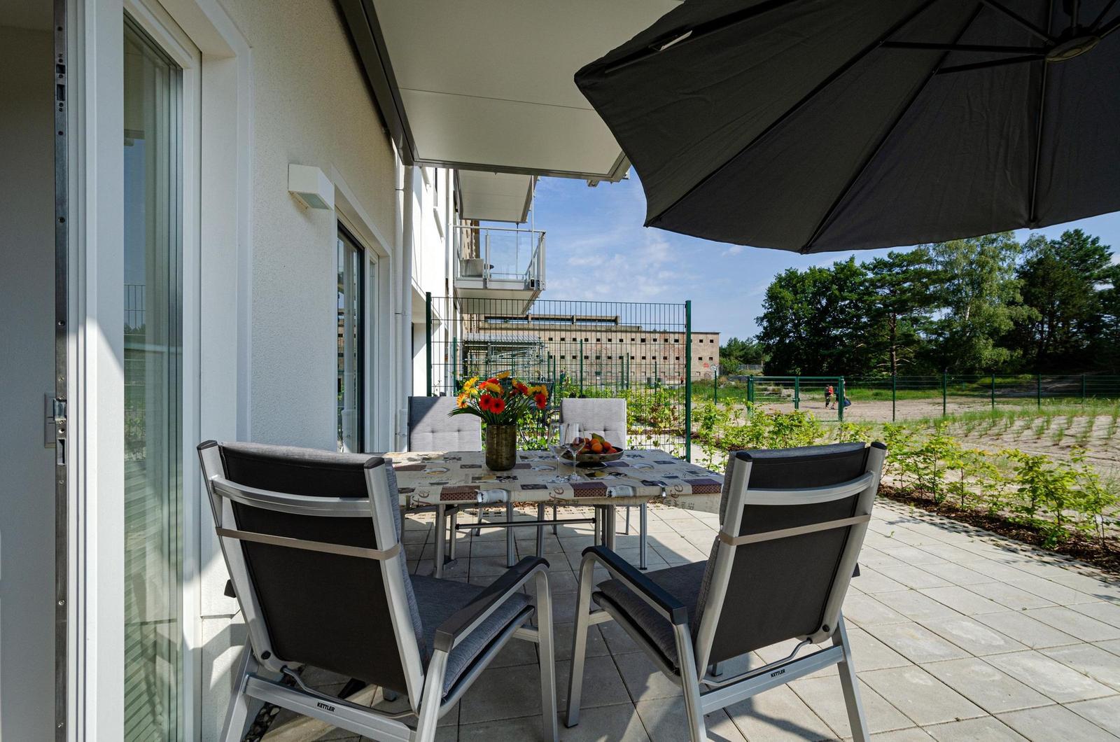 Terrasse mit Tisch, Stühlen und Sonnenschirm. Blick auf Garten und Gebäude.