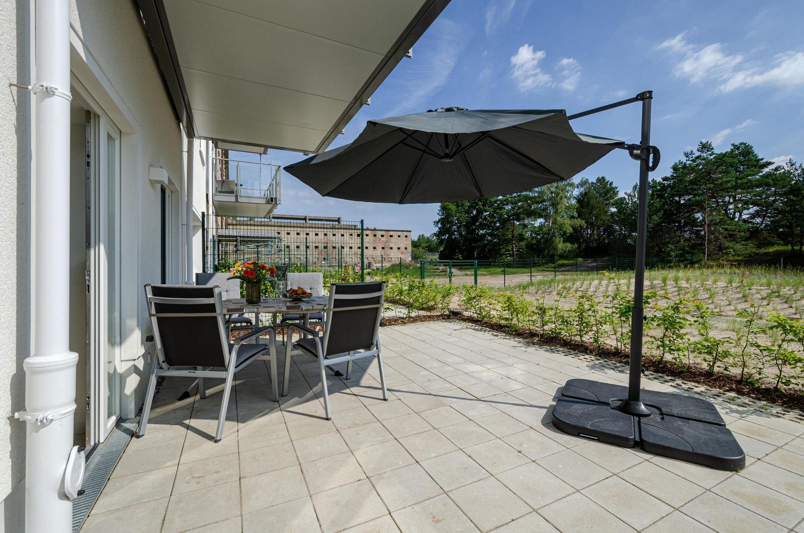 Terrasse mit Tisch, Stühlen und Sonnenschirm. Hintergrund: Grünfläche und Bäume.