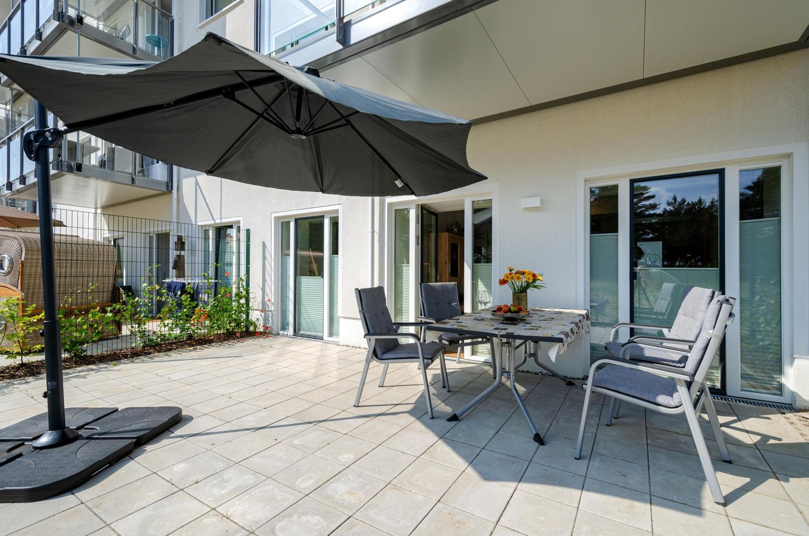 Terrasse mit Tisch, Stühlen und Sonnenschirm. Hintergrund: Haus mit Fenstern und Pflanzen.