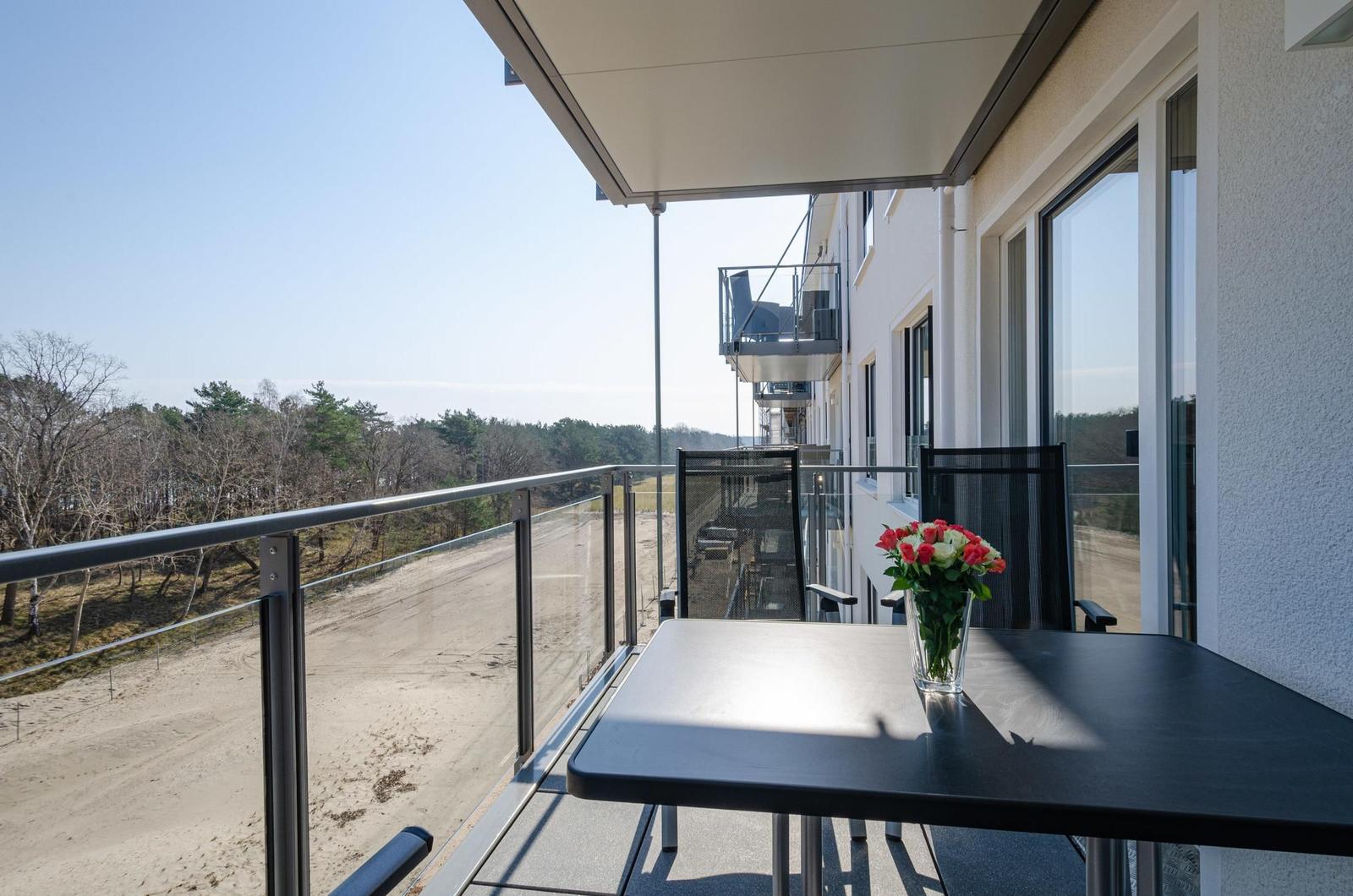 Balcony with table, chairs, and flowers. View of forest and sandy beach.