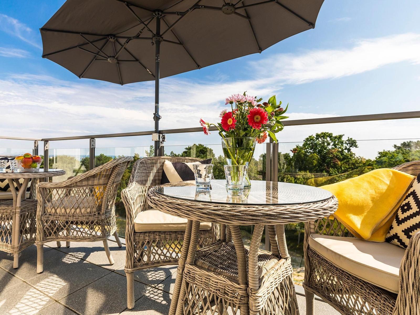 Terrasse mit Wickermöbeln, Sonnenschirm und Blumenarrangement. Blick auf grüne Landschaft.