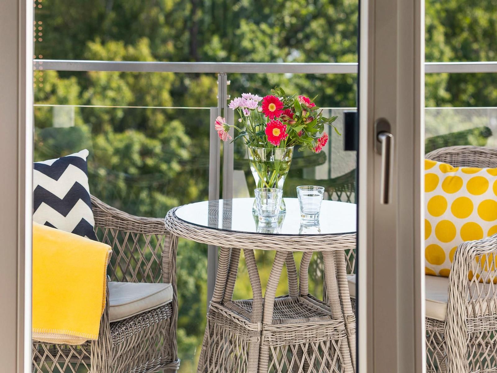 Terrasse mit Rattanmöbeln, Blumen und Gläsern. Blick auf grüne Bäume.