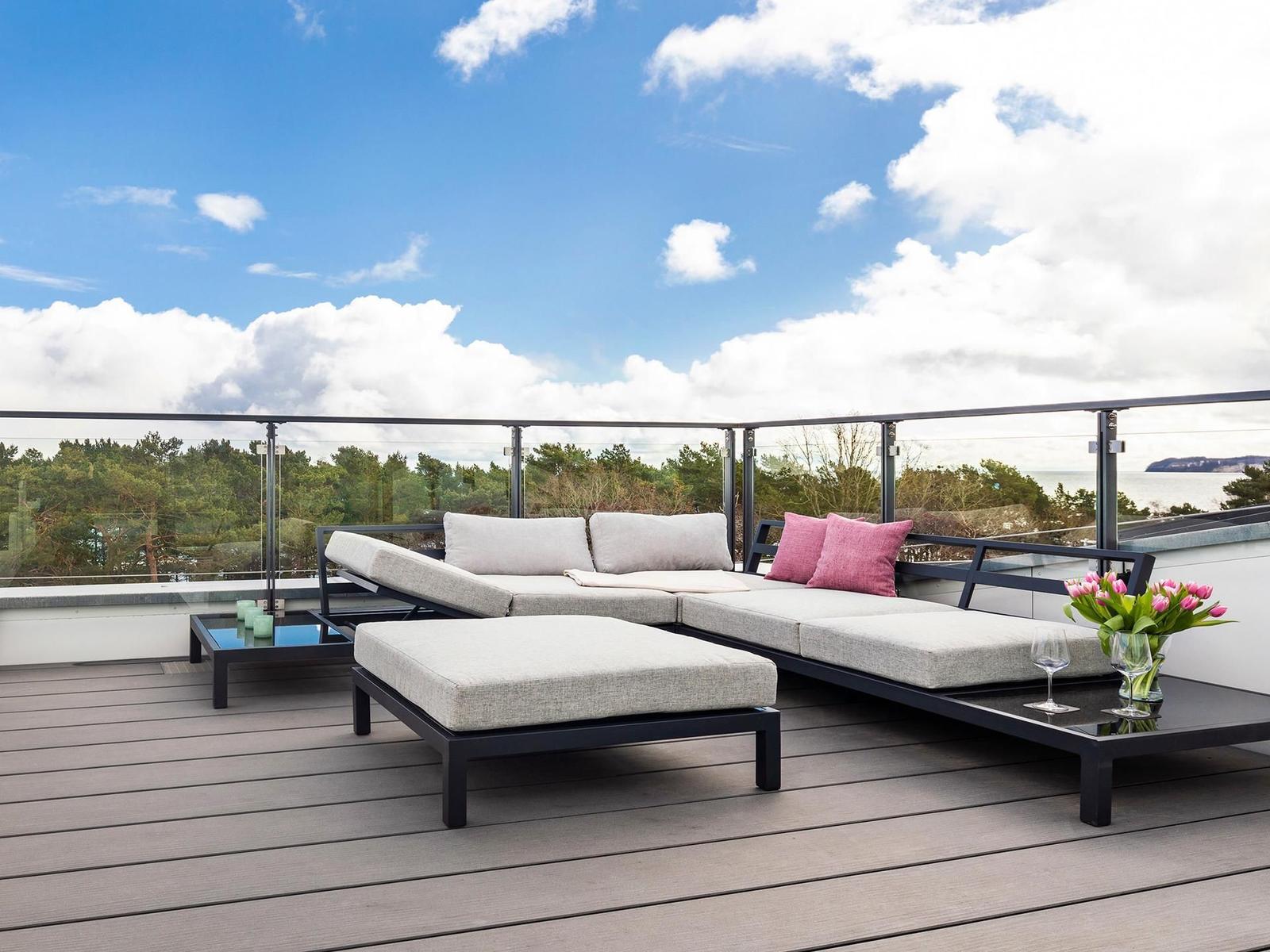 Terrasse mit Loungemöbeln, Blick auf Wald und Meer unter blauem Himmel mit weißen Wolken.