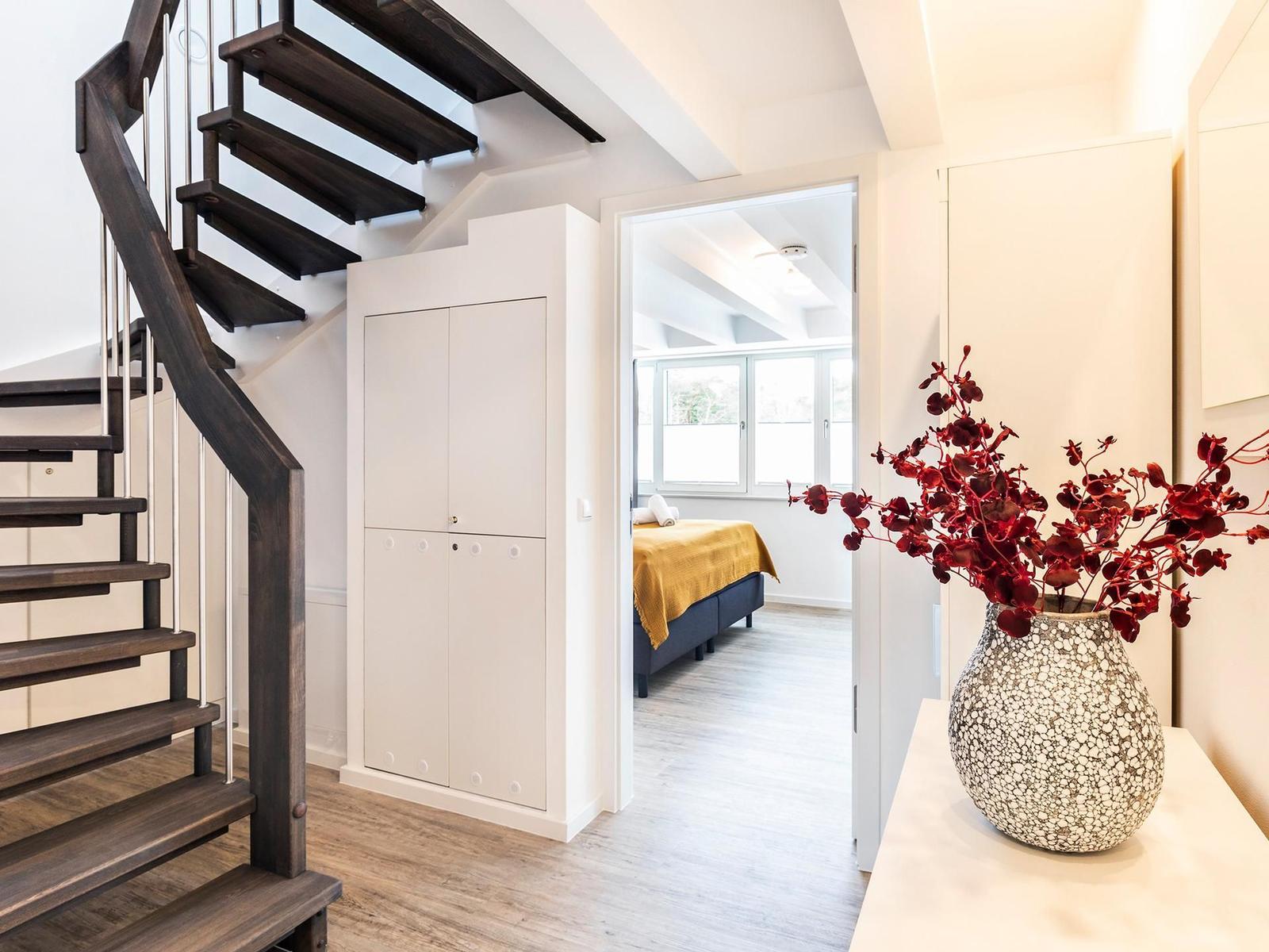 Eingangsbereich mit Treppe, Kleiderschrank und Blick in Schlafzimmer mit Bett und Fenster.