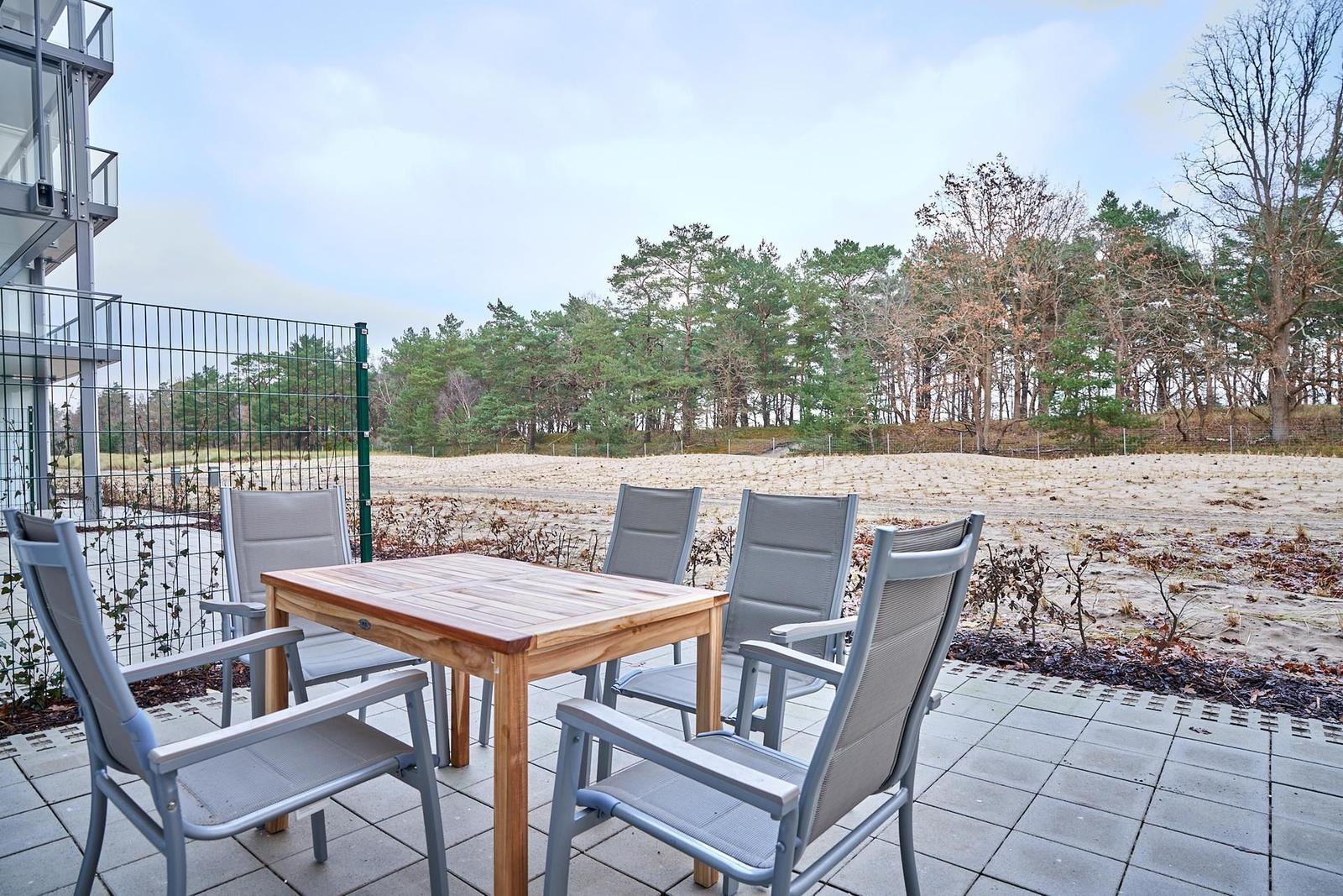 Terrasse mit Tisch und vier Stühlen, Blick auf Wald und Sandfläche.