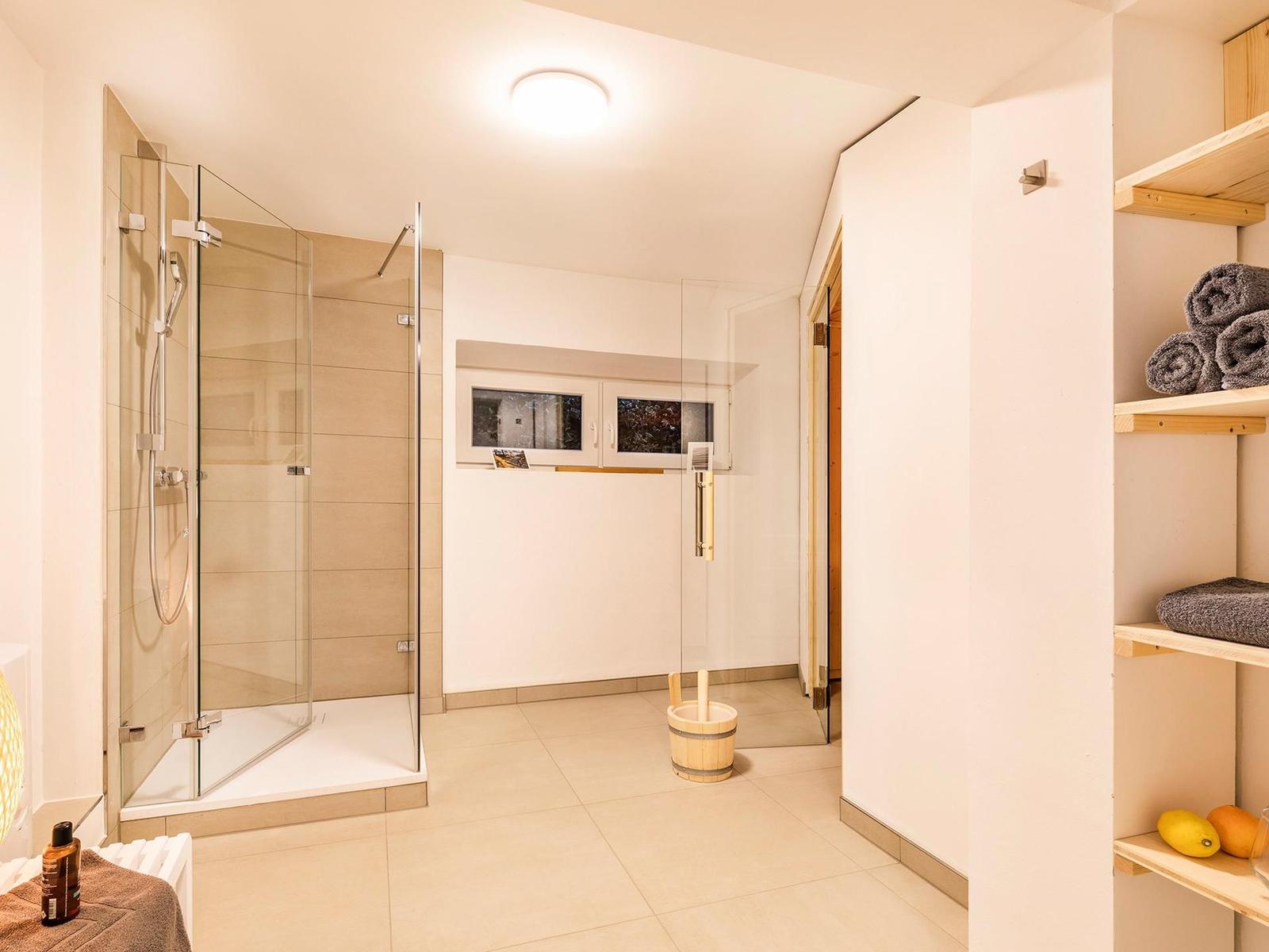 Dusche mit Glaswand, Sauna und Holzregale mit Handtüchern und Obst.