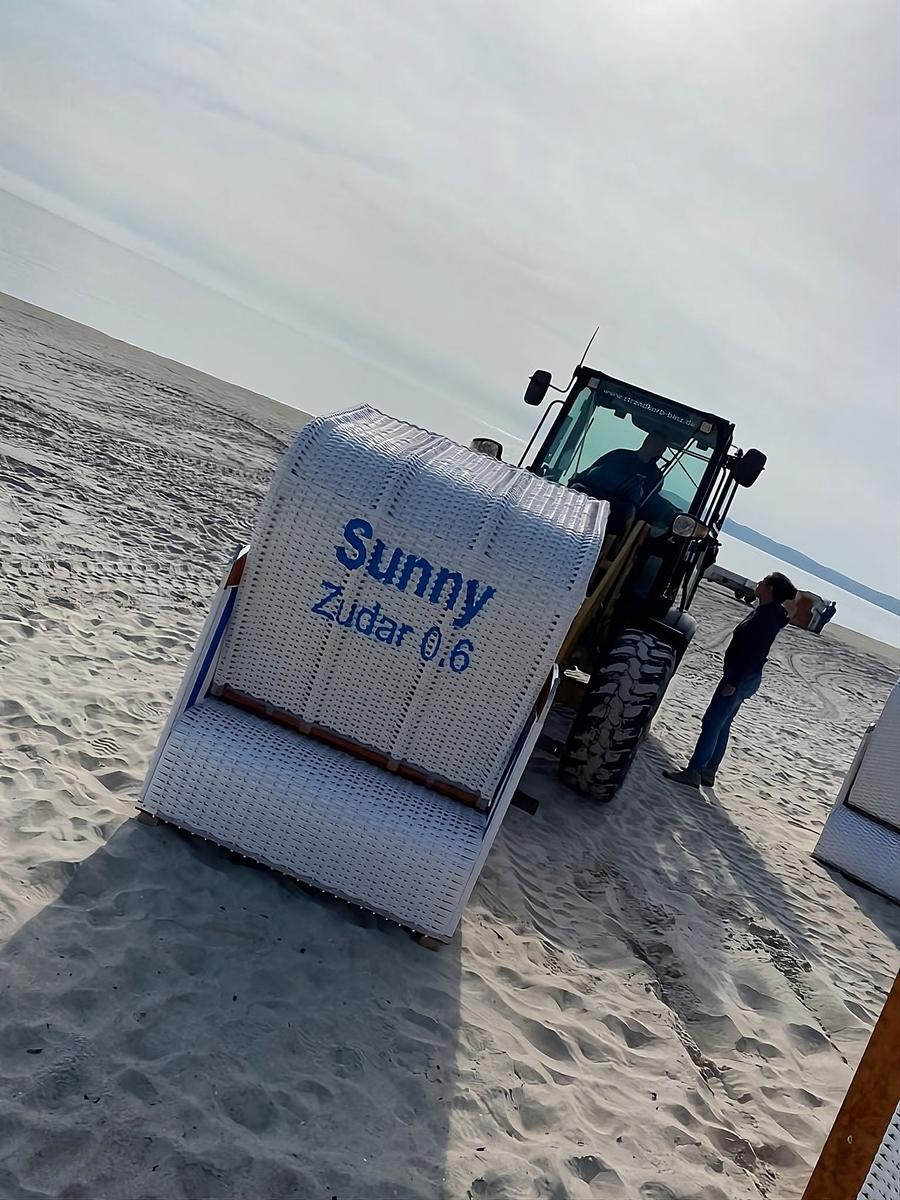 Weißer Strandkorb mit Aufschrift Sunny auf Sand, neben einem Traktor.