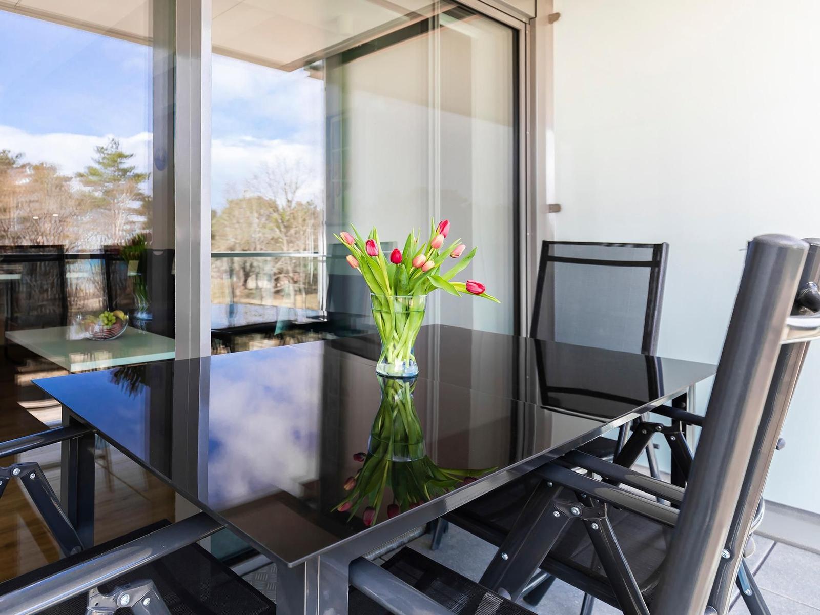 Terrasse mit Glas-Tisch, Stühlen und Tulpenblumen. Blick auf Bäume durch Glaswand.
