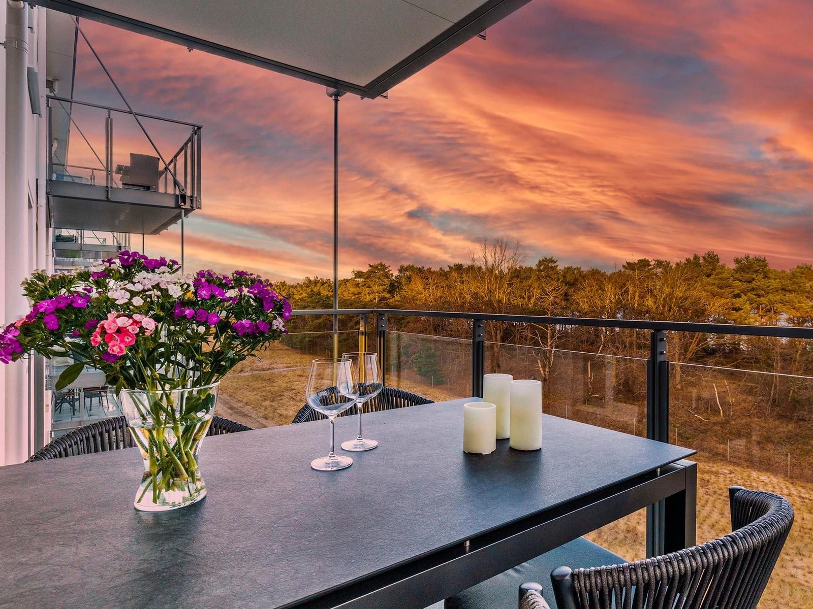 Terrasse mit Tisch, Stühlen, Blumen und Kerzen. Blick auf Wald bei Sonnenuntergang.