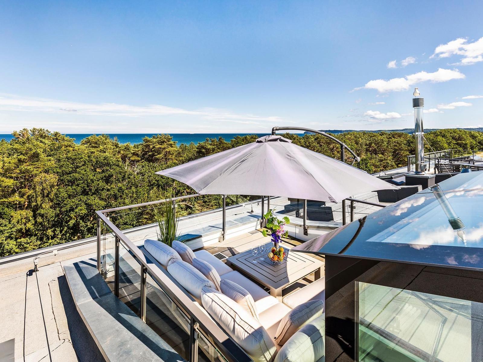 Terrasse mit Sitzgruppe, Sonnenschirm und Blick auf Meer und Wald.