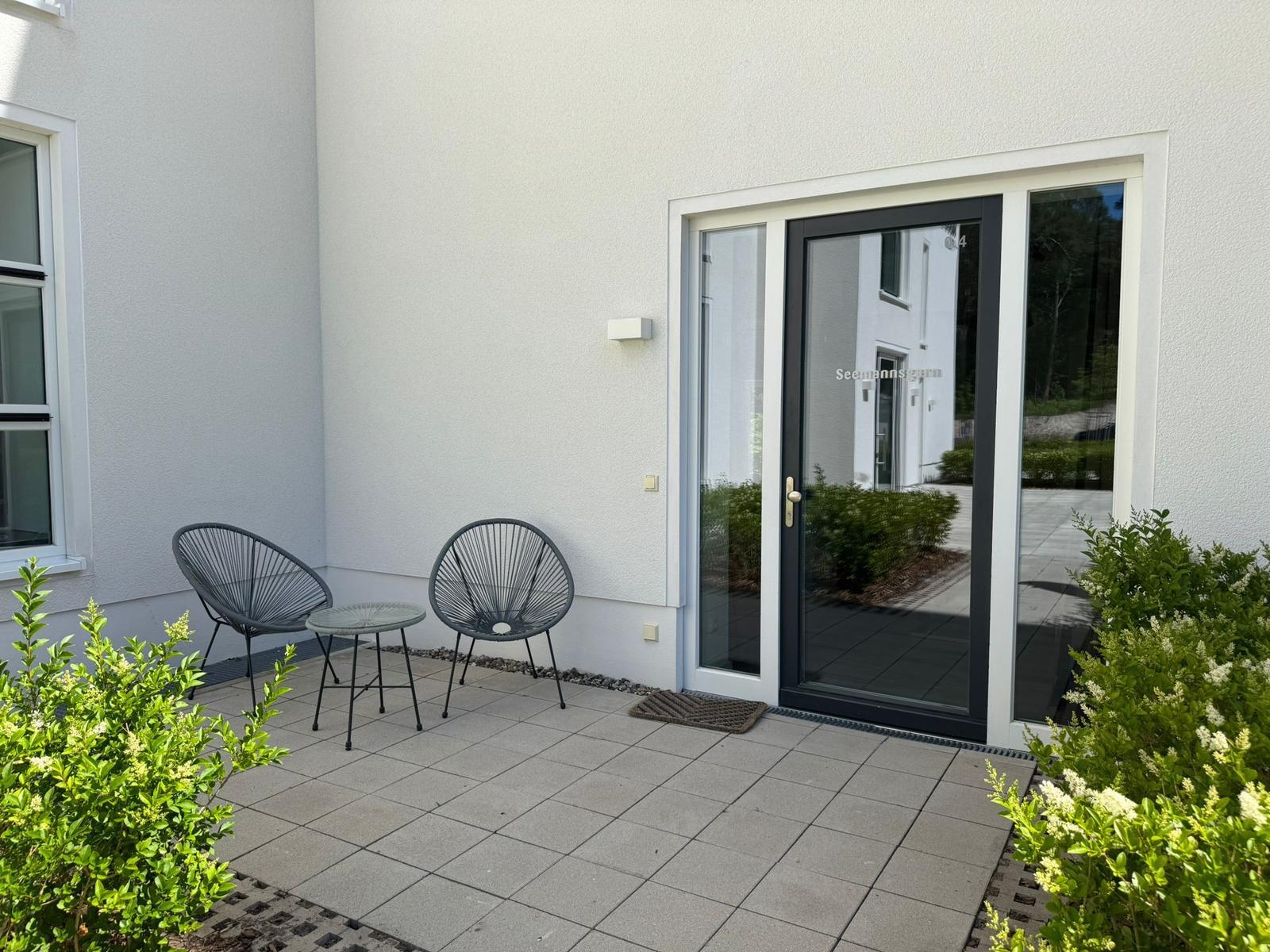 Terrace with two chairs and table in front of glass door. Surrounded by plants and light walls.