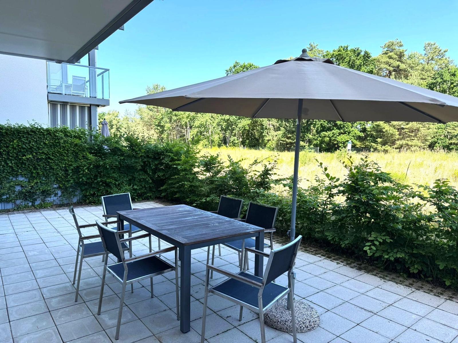 Terrace with table, four chairs, and umbrella. Background: grassy area and trees.