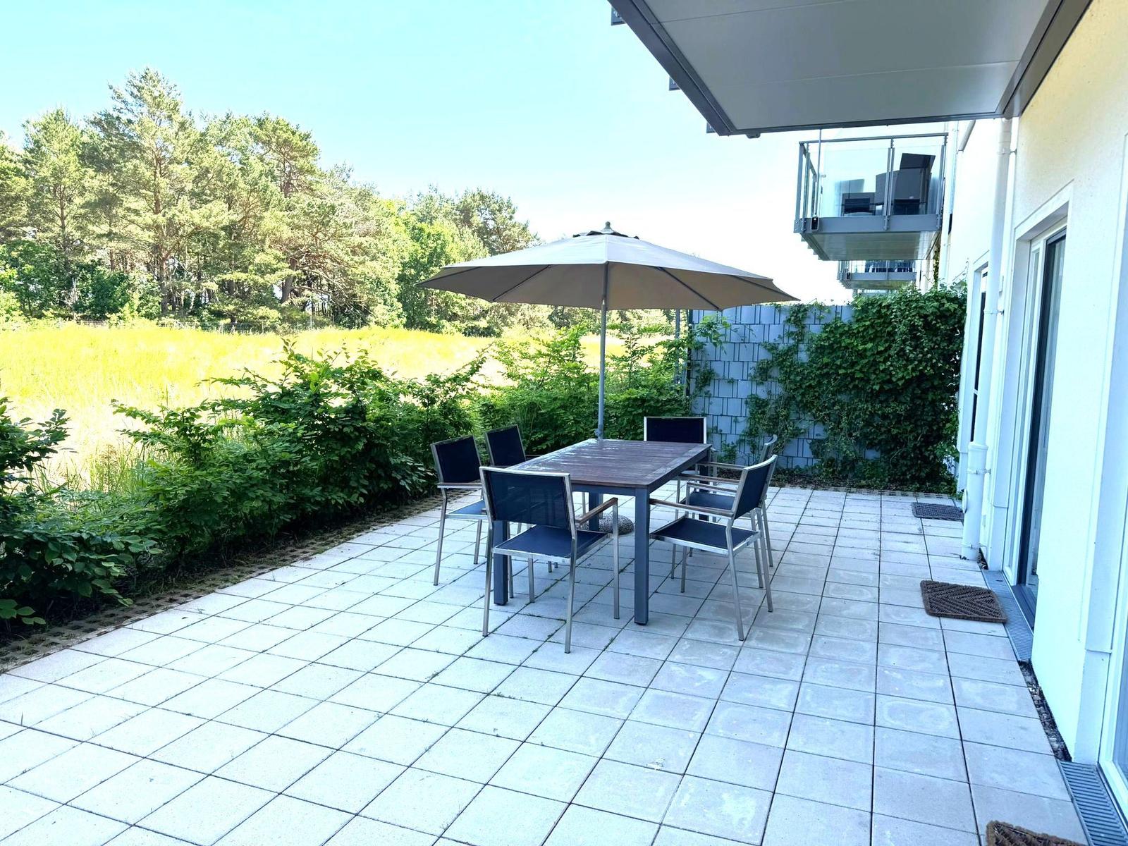 Terrace with table, chairs, and umbrella. Background: green field and trees.