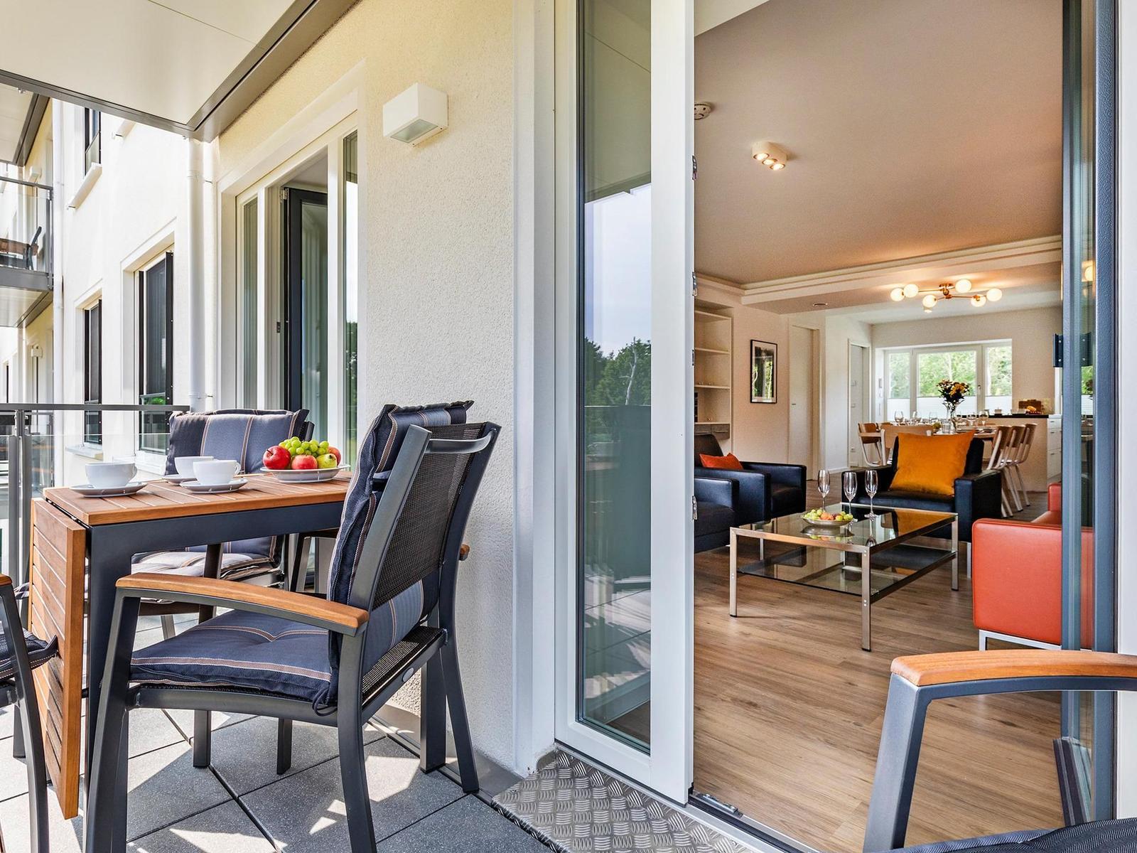 Terrace with table and chairs. Through glass doors, living room with sofa and couch is visible.