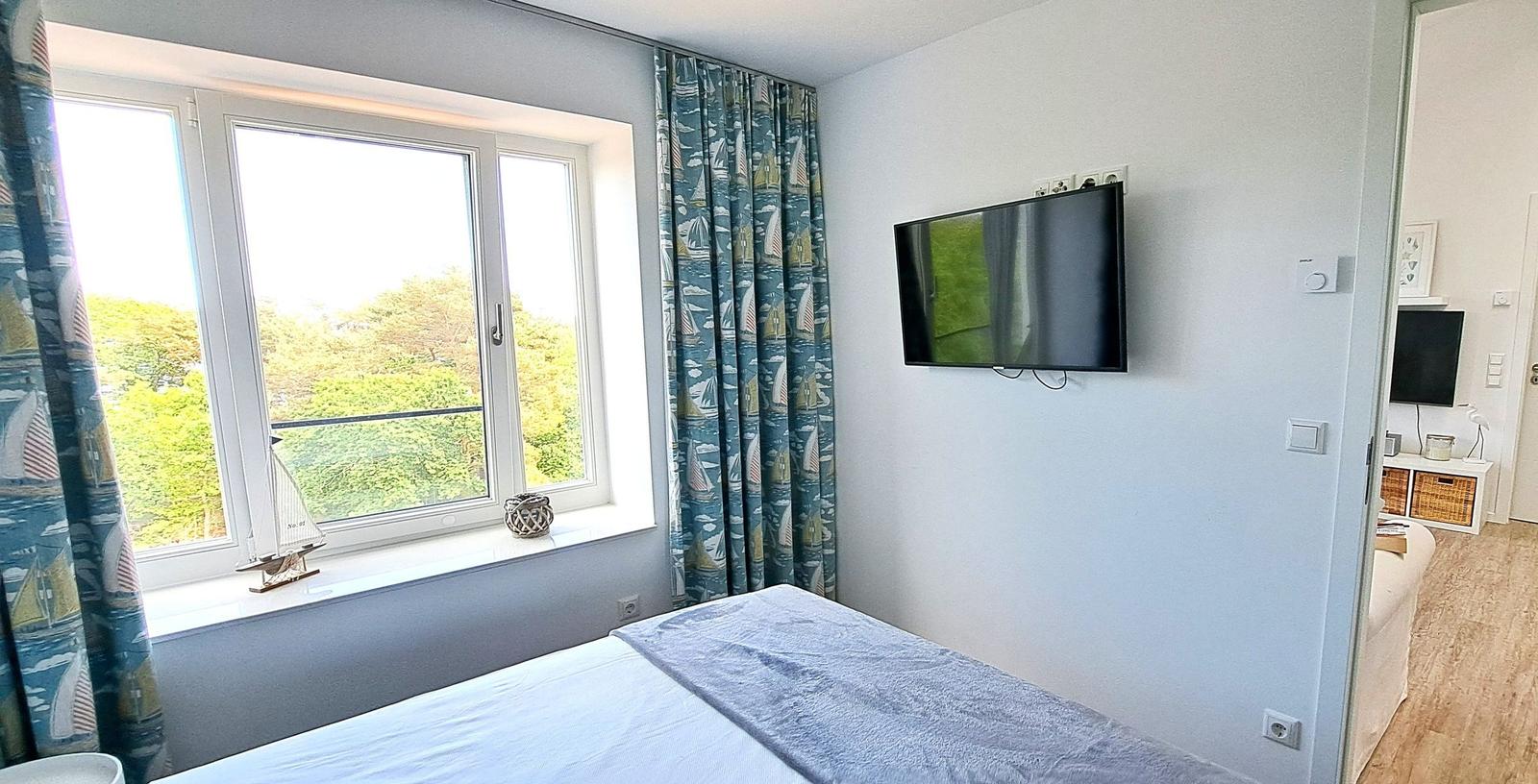 Bedroom with view of greenery, TV mounted on wall, bed with gray cover, sailboat-patterned curtains.
