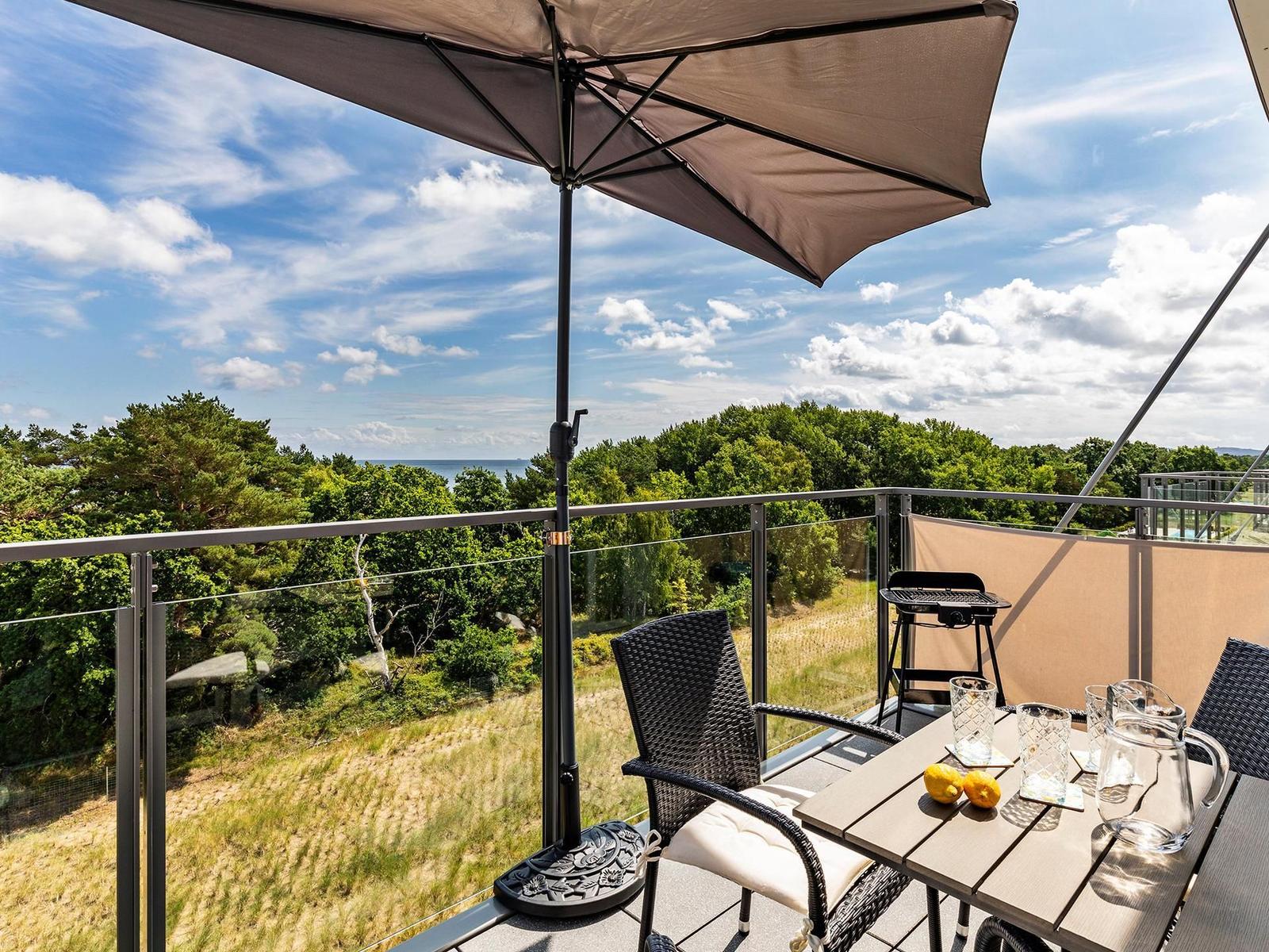 Terrace with table, chairs, and umbrella. View of forest and sea.