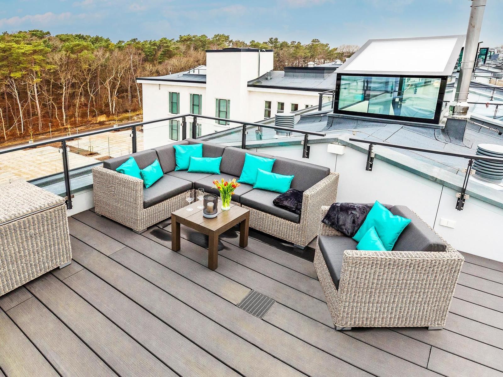 Terrasse mit Sofa, Sitzgruppe und Tisch. Blick auf Wald und Gebäude.