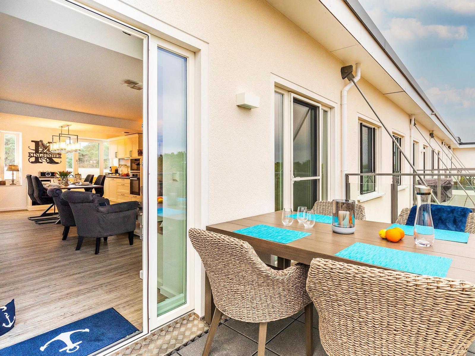 Terrasse mit Esstisch und Stühlen. Durch Glastür ist Wohn- und Essbereich sichtbar.