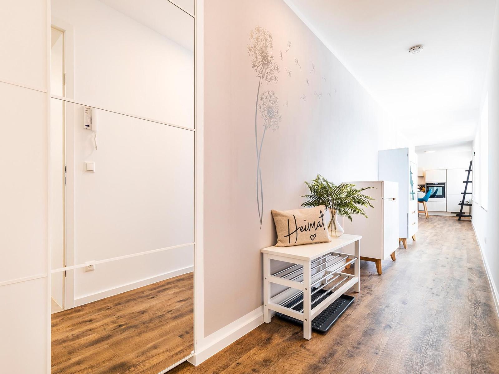 Eingangsbereich mit Schuhschrank, Pflanze und Wanddekoration. Holzboden und weißer Schrank sind sichtbar.