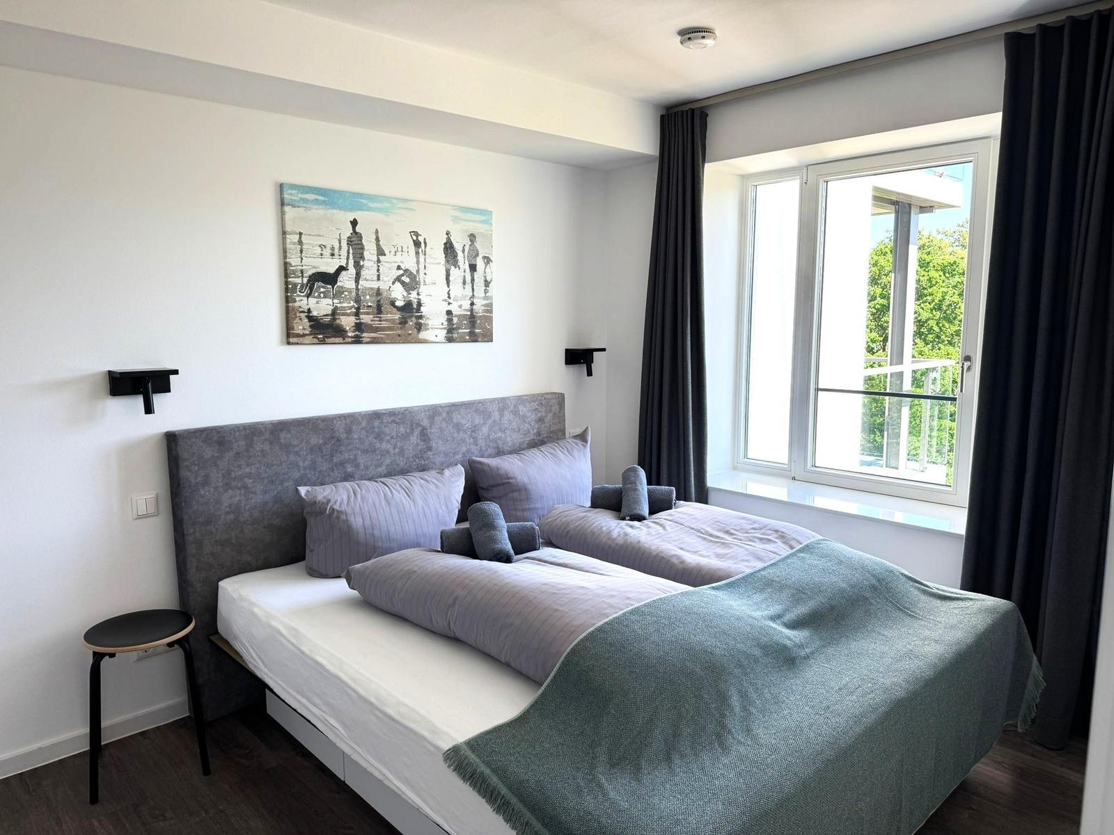 Schlafzimmer mit Doppelbett, Wandbild und großem Fenster mit Blick auf Balkon.