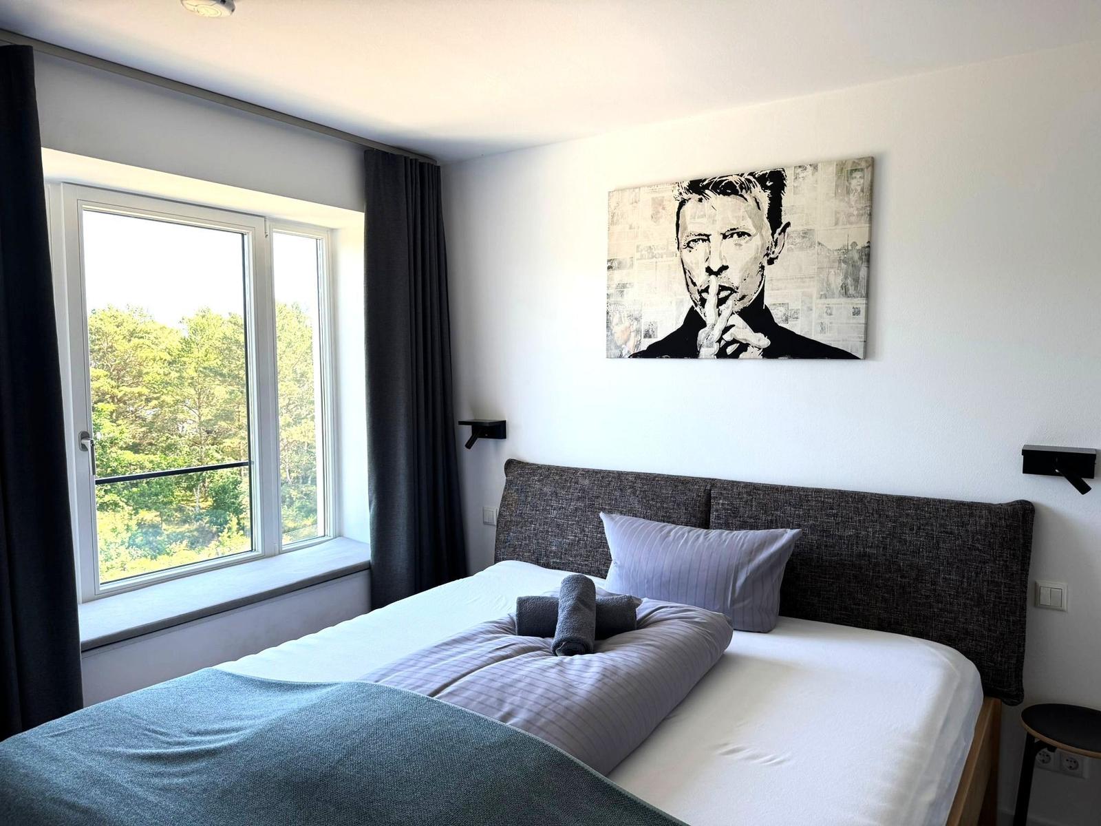 Schlafzimmer mit Bett, Fenster und Wandbild. Blick auf grüne Bäume.