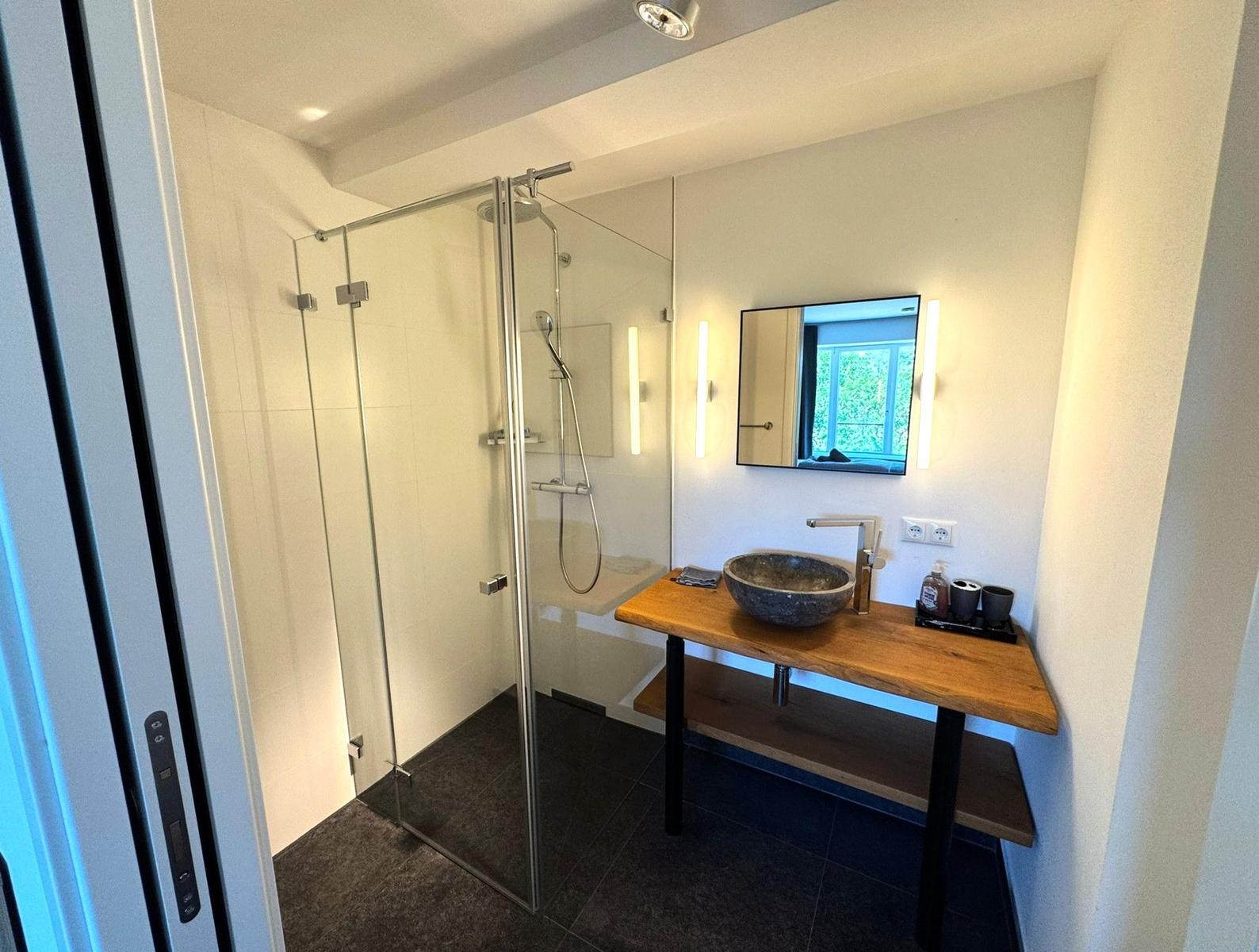 Dusche mit Glaswand, Waschtisch mit Steinwaschbecken und Spiegel mit Beleuchtung.
