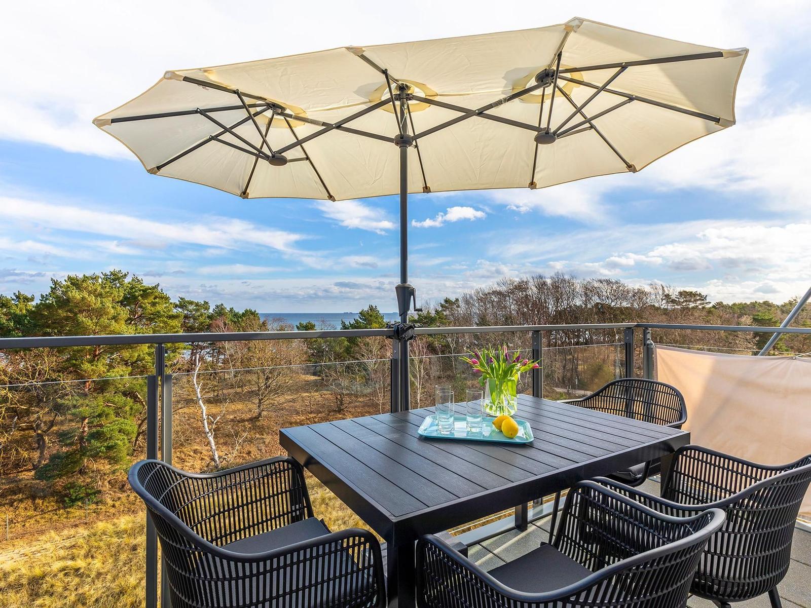 Terrasse mit Tisch, Stühlen und Sonnenschirm. Blick auf Wald und Meer.