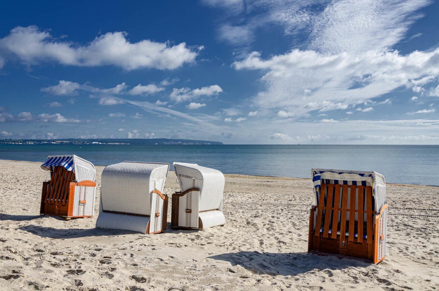 Drei Strandkörbe stehen am sandigen Strand unter blauem Himmel mit weißen Wolken.
