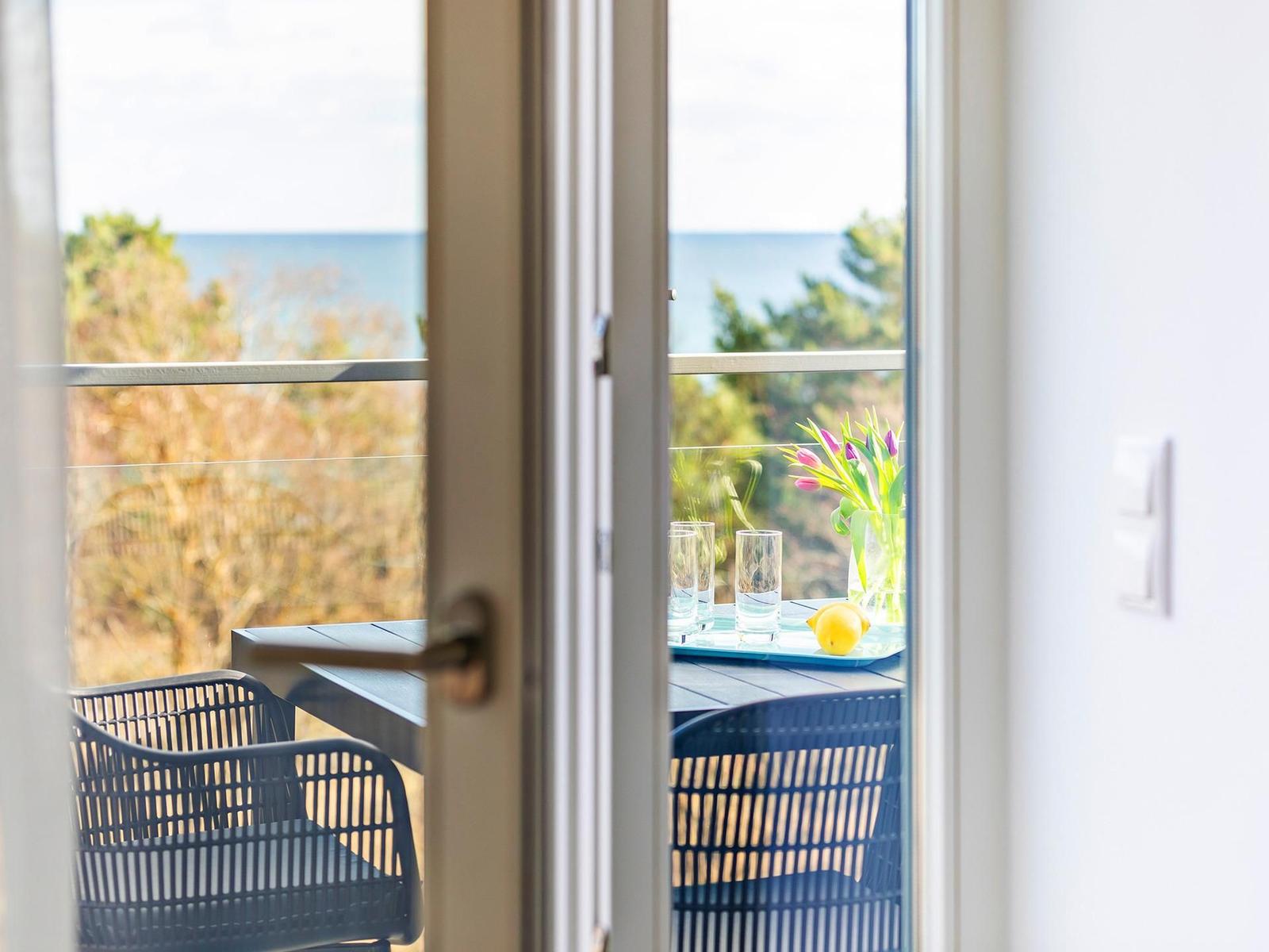 Durch die Tür sieht man einen Balkon mit Tisch, Stühlen und Blick auf Meer und Bäume.