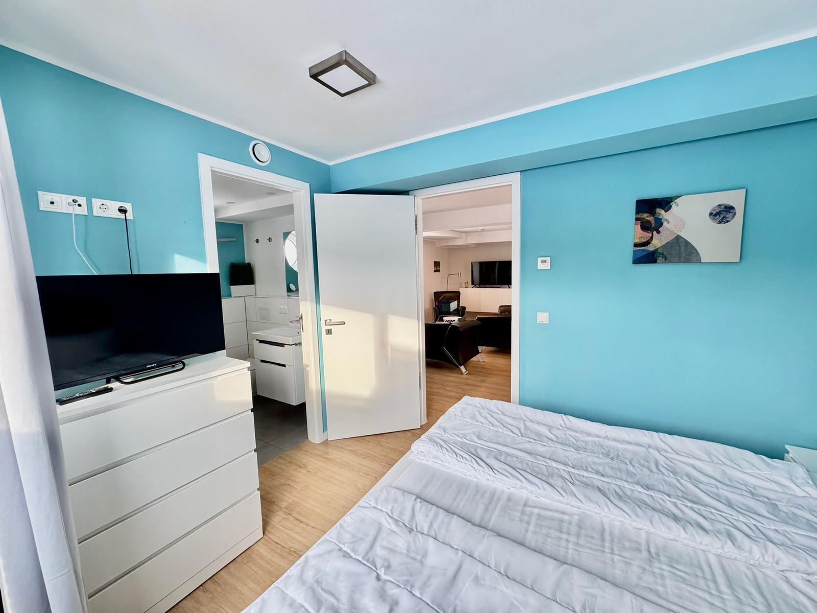 Schlafzimmer mit blauen Wänden, Bett, Fernseher und Blick in Bad und Wohnbereich.