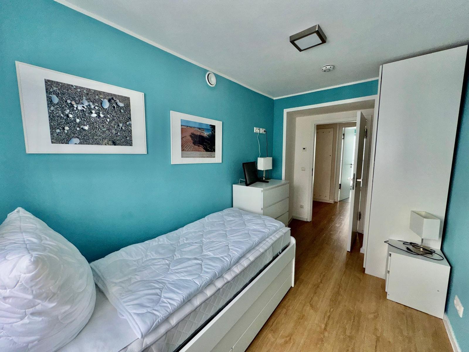 Schlafzimmer mit blauen Wänden, weißem Bett und Holzboden. Zwei Bilder an der Wand.