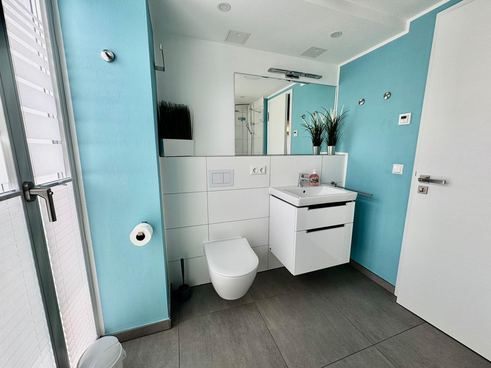 Badezimmer mit blauen Wänden, weißem Waschbecken und Toilette. Fenster mit Jalousie.