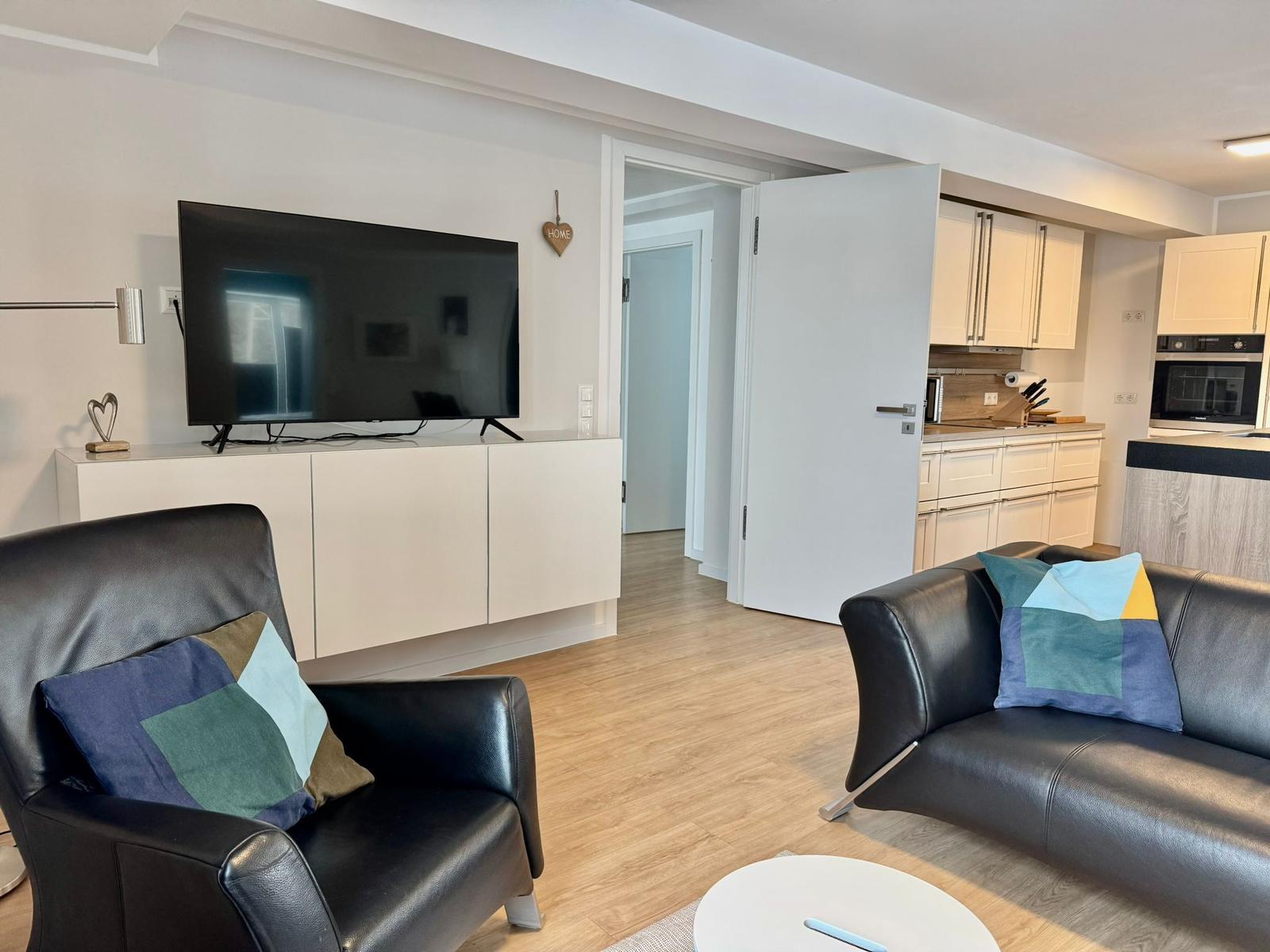 Wohnzimmer mit Sofa, Sessel und TV. Küche ist im Hintergrund sichtbar.