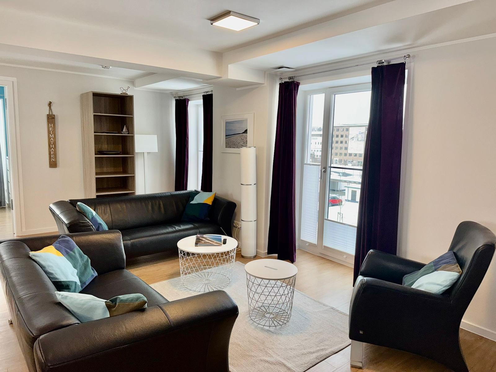 Wohnzimmer mit Ledersofa, Sessel und zwei runden Tischen. Dunkelviolette Vorhänge an Fenstern.