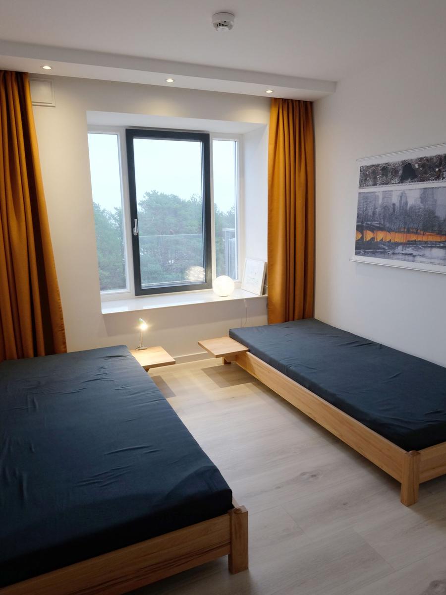 Zwei Holzbetten mit dunklen Matratzen. Fenster mit orangefarbenen Vorhängen und Wandbild.