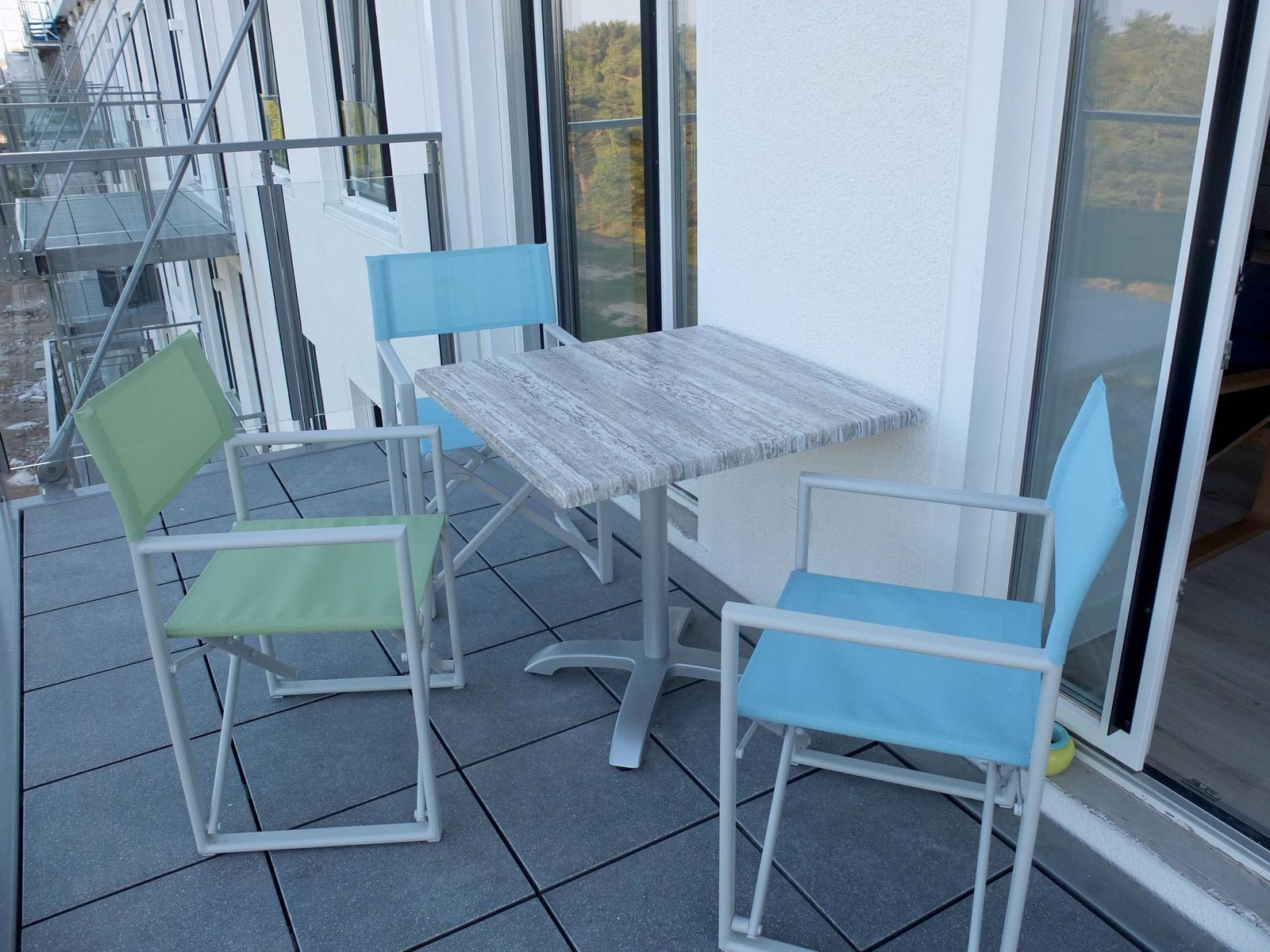 Terrasse mit Tisch und drei Stühlen, zwei blau, einer grün, Blick auf Wald.