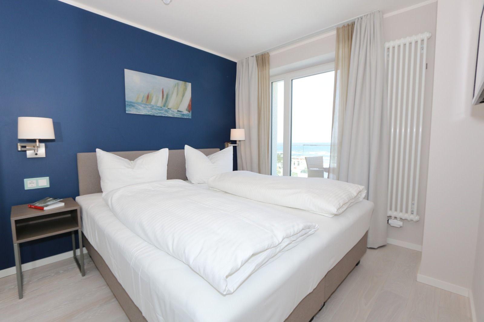 Schlafzimmer mit Doppelbett, blauer Wand und Meerblick durch Fenster.