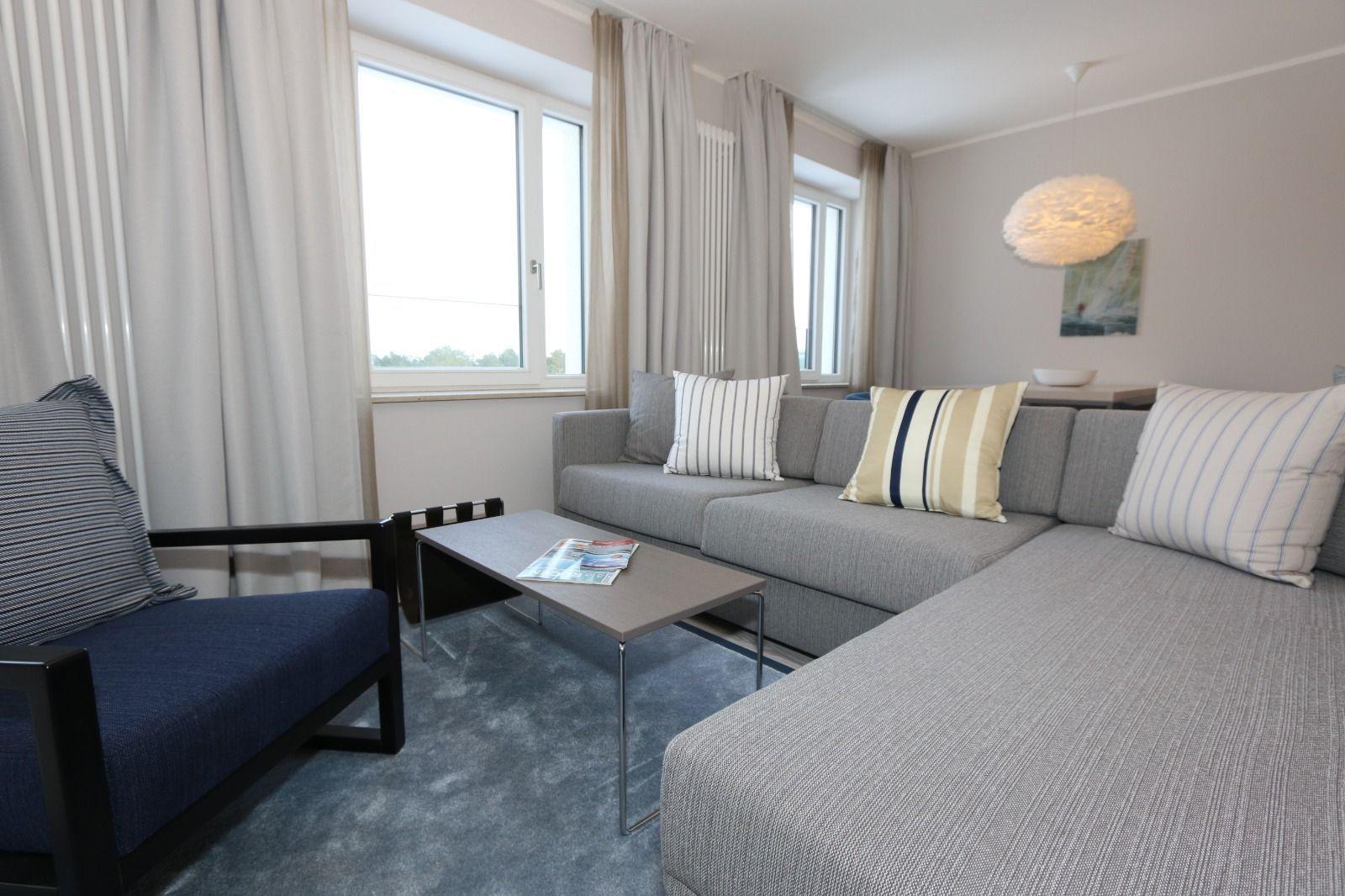 Wohnzimmer mit Sofa, Sessel, Couchtisch und Fenstern mit Vorhängen.