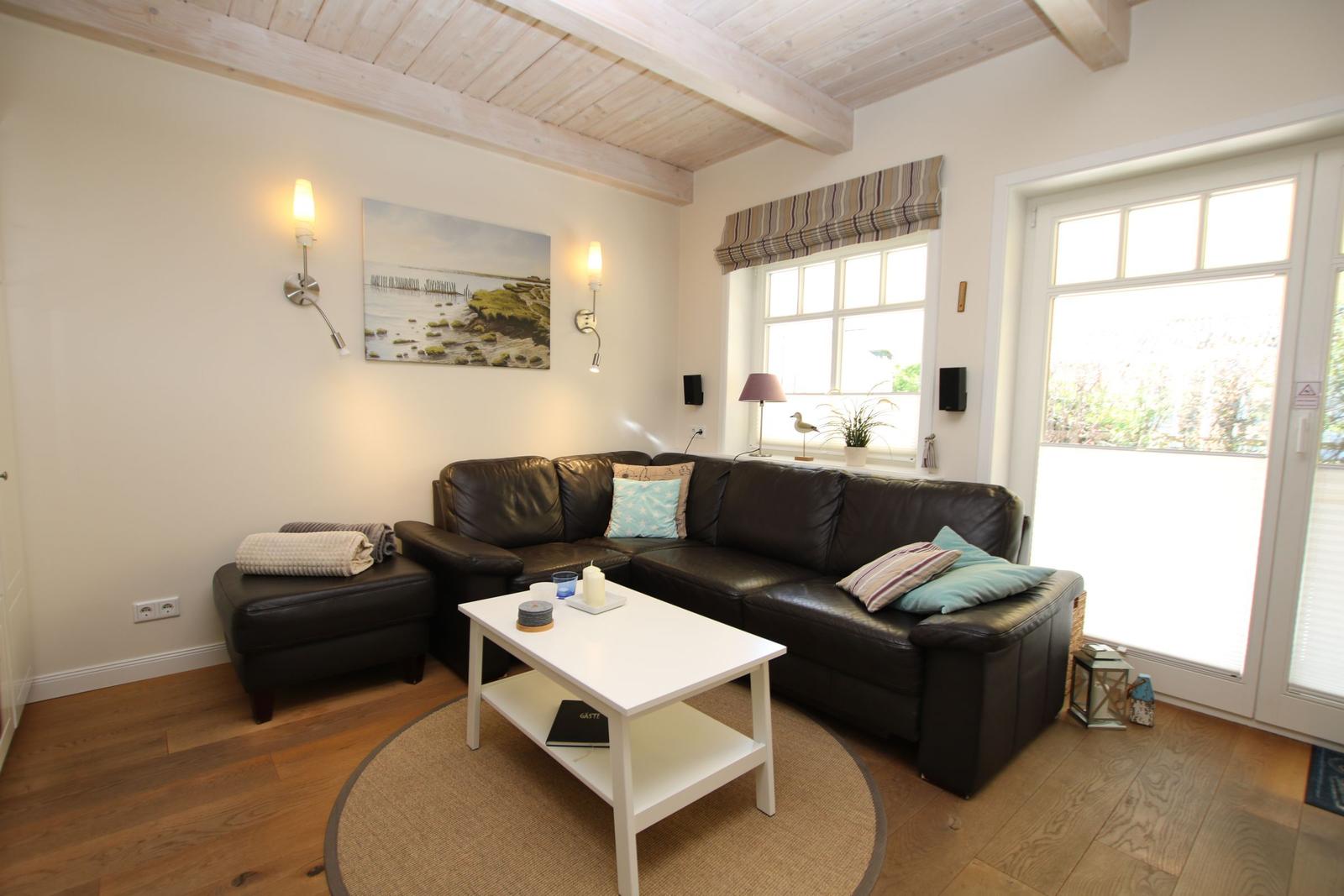 Wohnzimmer mit braunem Sofa, weißem Tisch und Holzboden. Fenster und Tür mit Blick nach draußen.