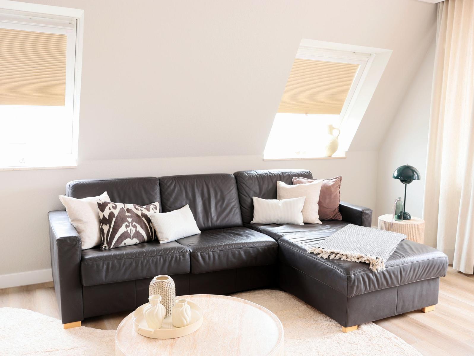 Schwarzes Ledersofa mit Kissen und Decke, runder Tisch, Fenster mit Jalousien.