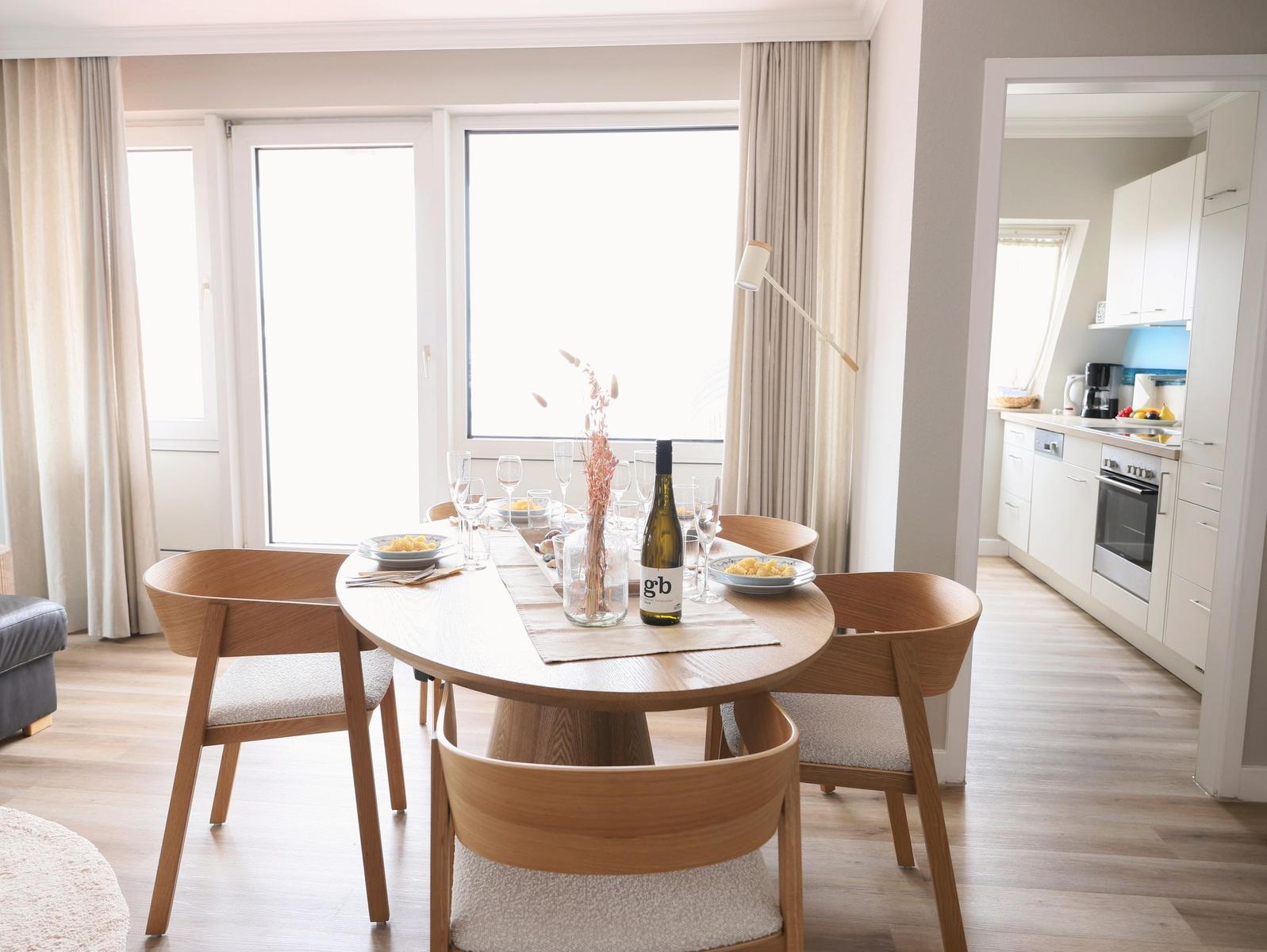 Esszimmer mit Holztisch, Stühlen und Fenster. Küche im Hintergrund sichtbar.