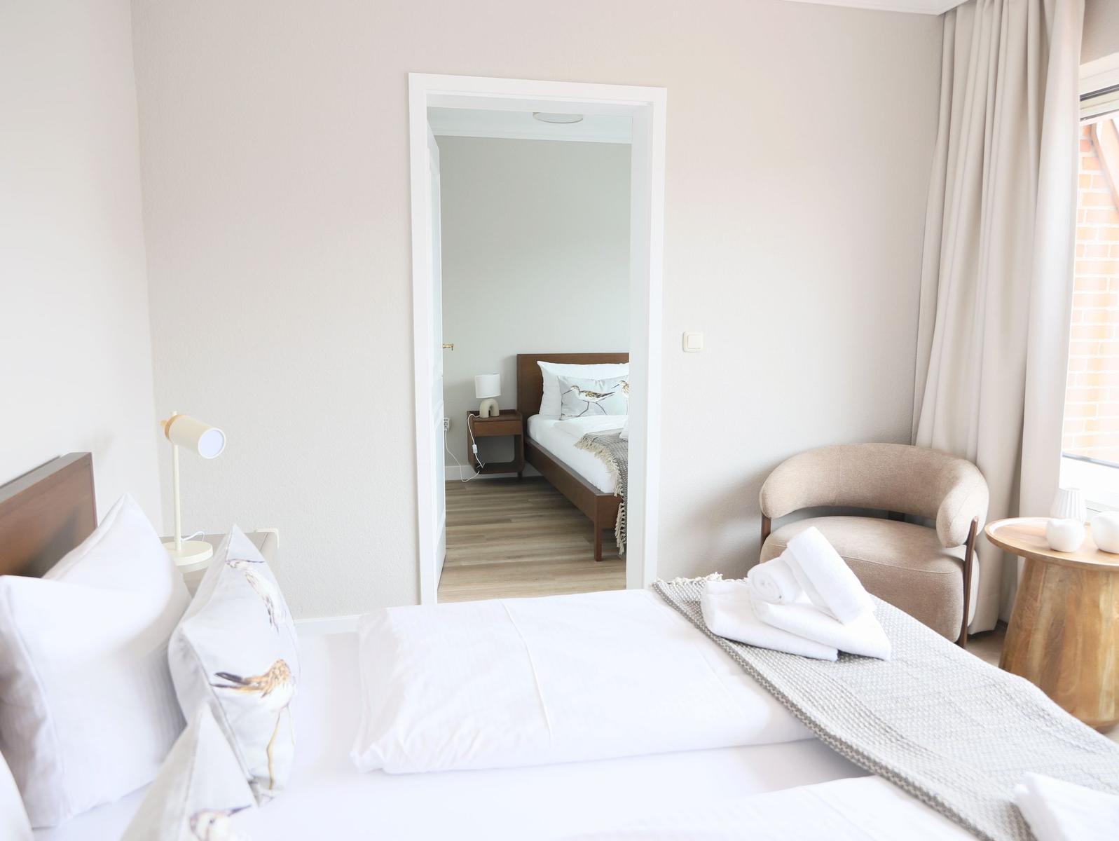 Schlafzimmer mit Bett, Stuhl und Fenster. Durchgang zu einem weiteren Schlafzimmer.