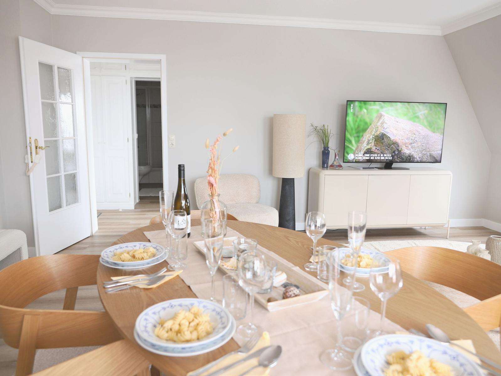 Gesetzter Tisch mit Tellern und Gläsern. Fernseher und Sessel im Hintergrund.
