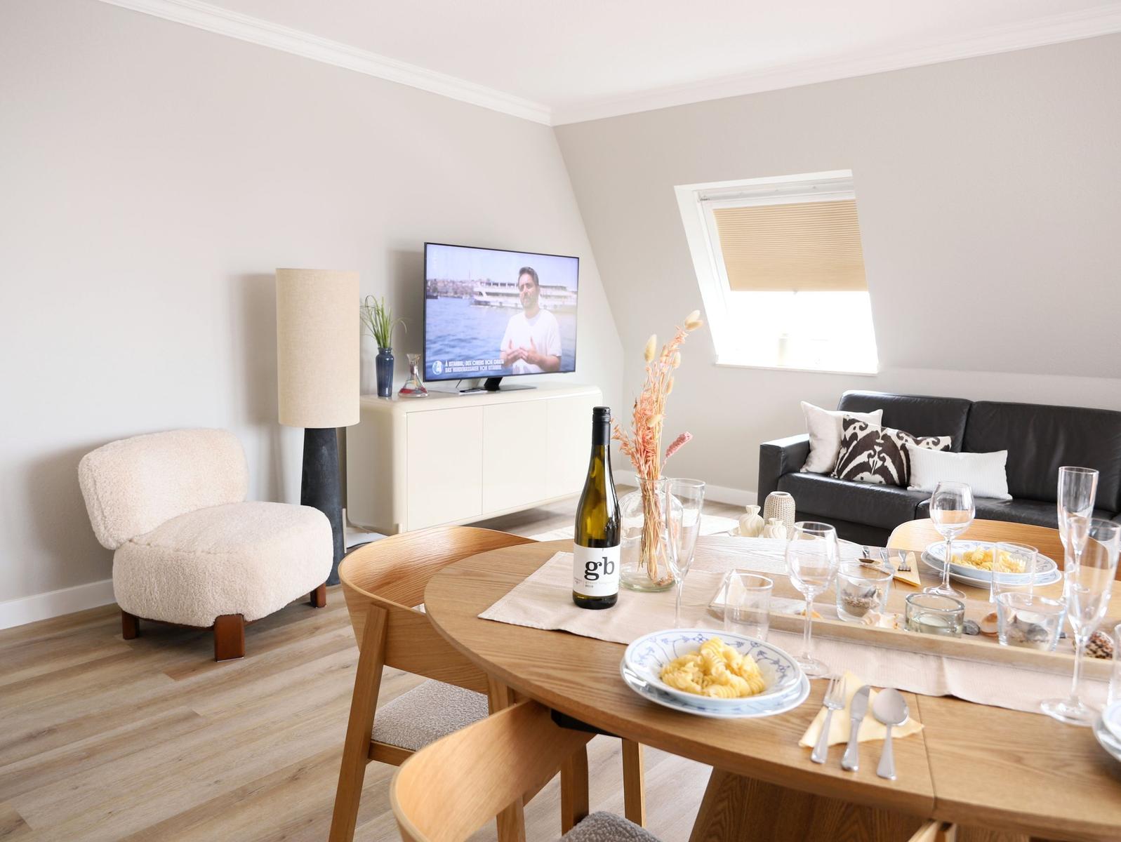 Esszimmer mit Holztisch, Sofa und Fernseher. Auf dem Tisch stehen Gläser und eine Flasche.