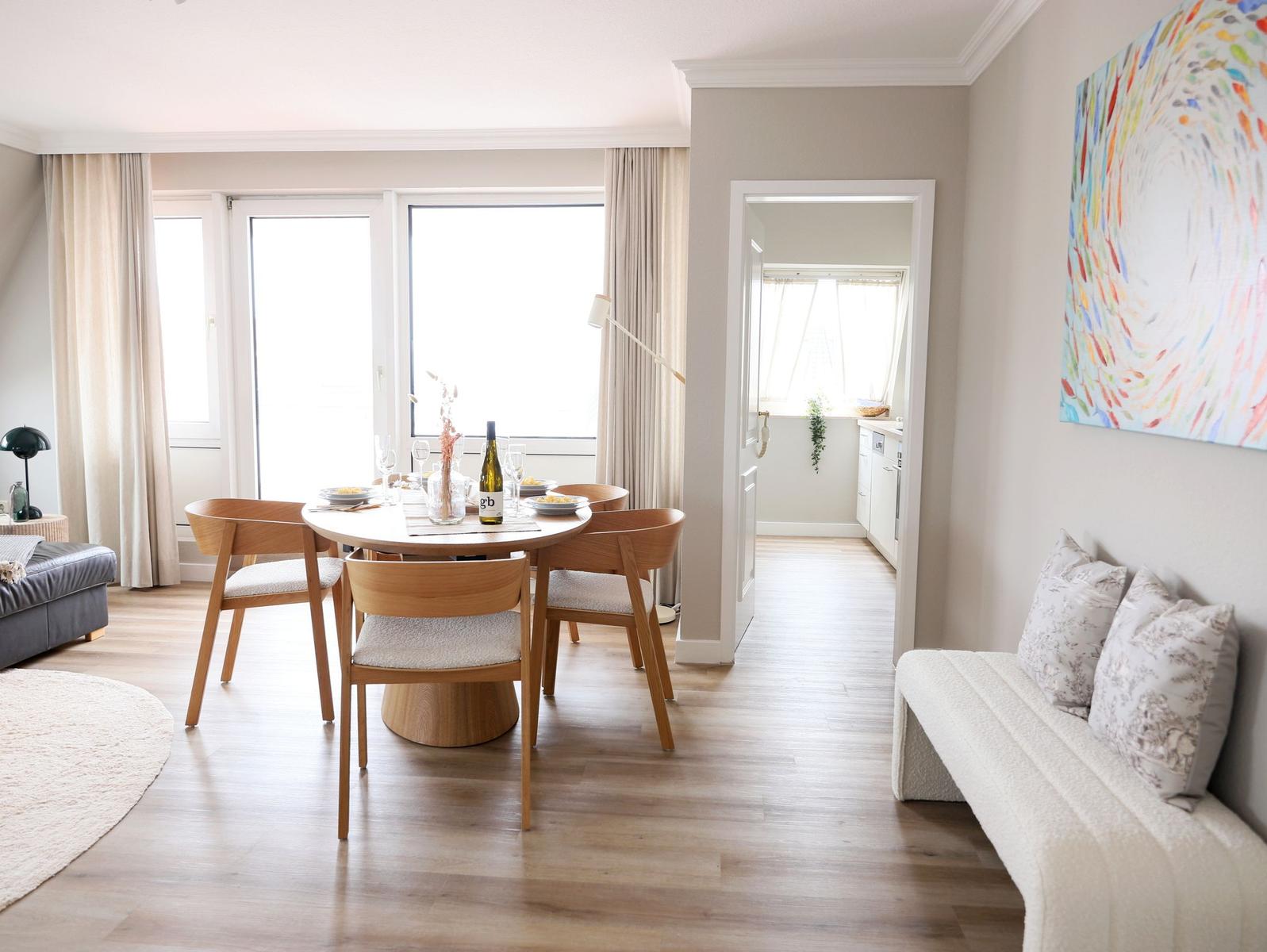 Esszimmer mit Tisch, Stühlen und Blick auf Balkon. Kuche im Hintergrund. Wandbild und Sofa sichtbar.