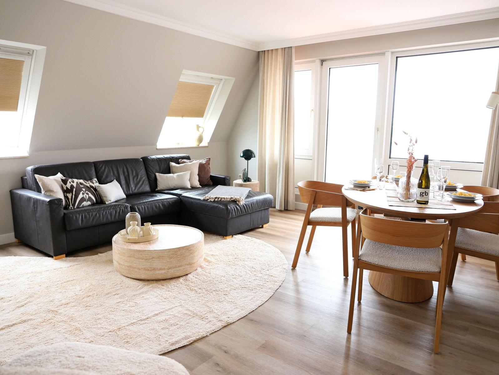 Wohn- und Essbereich mit Sofa, Tisch und Stühlen. Holzboden und große Fenster.