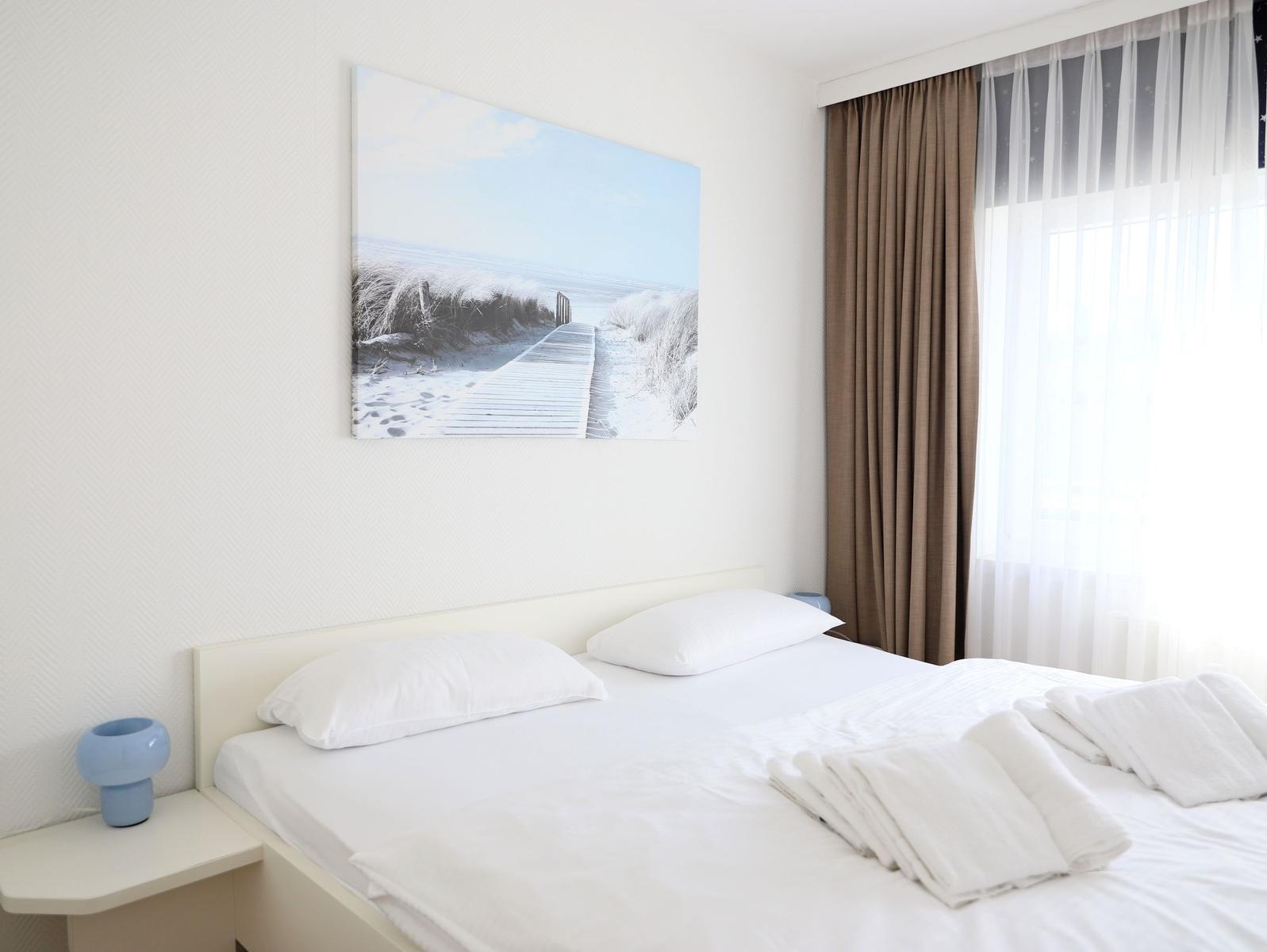 A bed with white linens and towels. Wall art shows a beach. Curtains are brown and white.