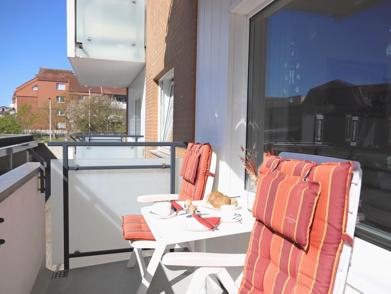 Terrace with table, two chairs, and orange cushions. Breakfast set and view of buildings.