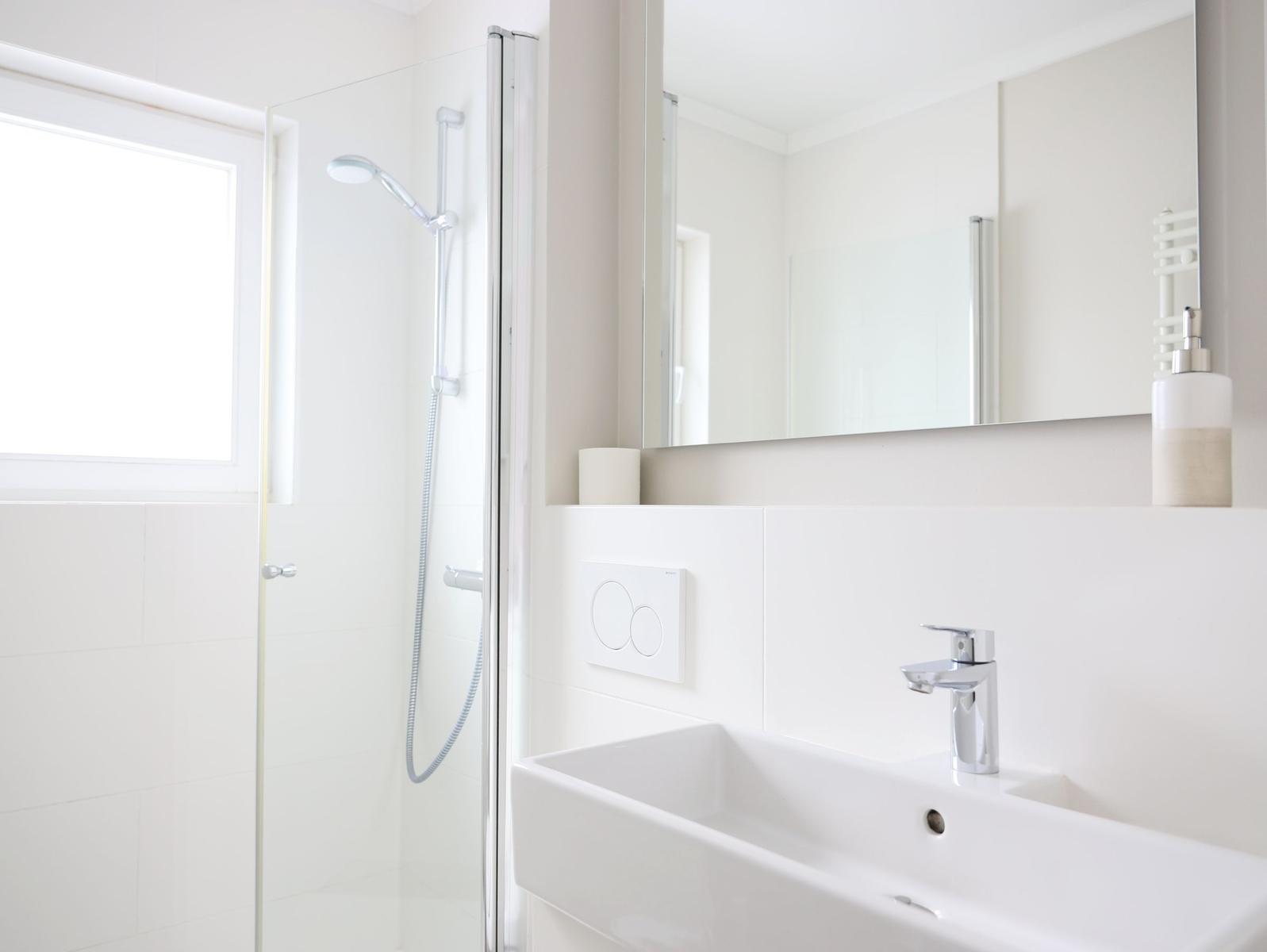 Badezimmer mit Dusche, Waschbecken und Spiegel. Fenster sorgt für Tageslicht.